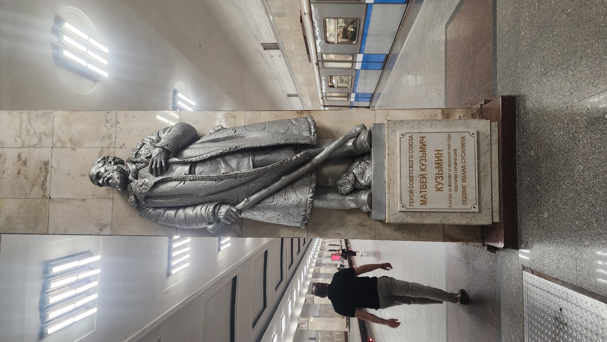 This image captures a vibrant scene inside a Moscow Metro station, showcasing a prominent monument alongside daily commuter activity. The central subject is a large, grey-colored relief statue of an elderly man, likely metallic, mounted on a marble-tiled wall. He is depicted wearing a long coat and holding a staff or walking stick, with a determined expression.
Below the statue, a white marble plaque with Cyrillic text provides context. The text reads: "ГЕРОЙ СОВЕТСКОГО СОЮЗА МАТВЕЙ КУЗЬМИЧ КУЗЬМИН В БИТВЕ ЗА МОСКВУ 14 ФЕВРАЛЯ 1942 ГОДА ПОВТОРИЛ ГЕРОИЧЕСКИЙ ПОДВИГ ИВАНА СУСАНИНА," which translates to "HERO OF THE SOVIET UNION MATVEY KUZMICH KUZMIN IN THE BATTLE FOR MOSCOW ON FEBRUARY 14, 1942, REPEATED THE HEROIC FEAT OF IVAN SUSANIN."
The setting is a well-lit subway platform. In the background, a blue and white subway train is visible on the tracks, indicating its presence at the station. The platform features polished grey flooring and white marble wall panels. Overhead, distinctive circular light fixtures with multiple horizontal fluorescent tubes illuminate the space.
A person wearing a black t-shirt and grey pants is walking away from the camera on the platform, while another figure can be seen further down. The image is taken at an angle, making the statue and the train appear somewhat tilted, but clearly shows the architectural grandeur often associated with Moscow's historic metro stations. The scene depicts a blend of historical commemoration and modern urban transit.