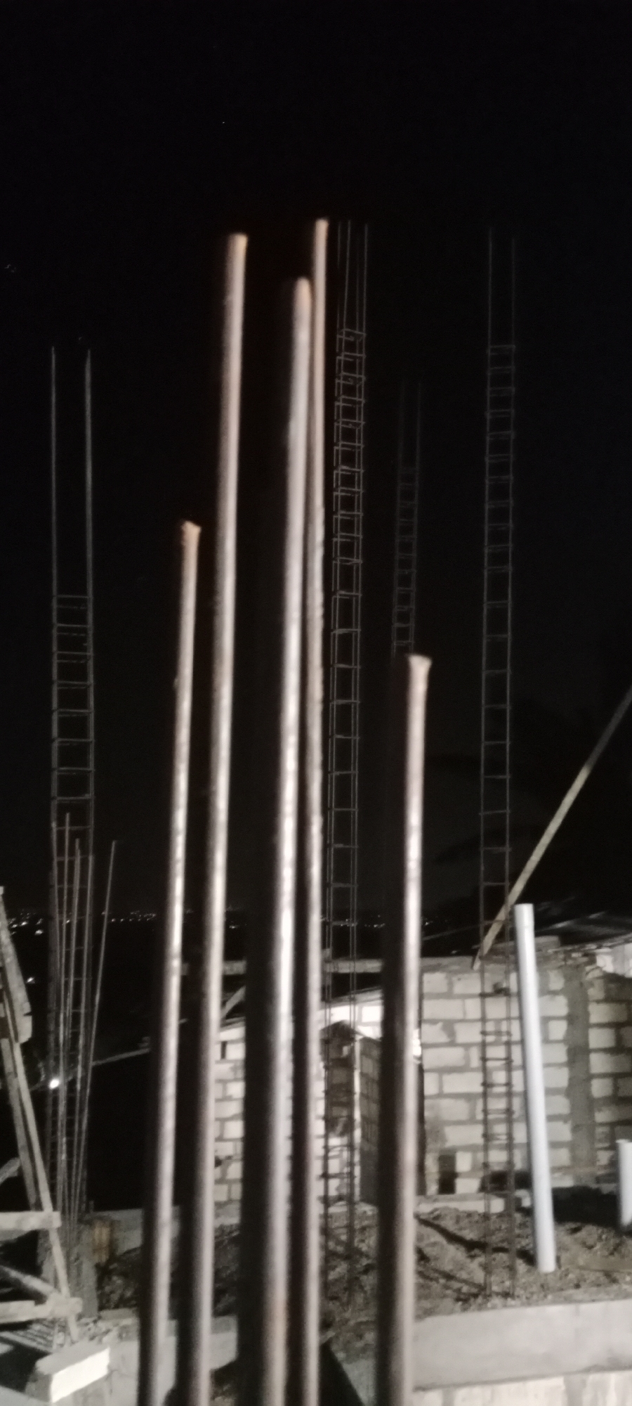 The image shows a construction site at night, possibly in Soko, Indonesia, given the context. The foreground is dominated by several thick, vertical rebar rods, some of which are partially illuminated. Behind these rods, the scene reveals more construction elements.
To the left, there are at least two tall, thin structures resembling ladder-like scaffolding, also made of rebar, rising into the darkness. In the mid-ground, more rebar columns are visible, some encased in what appears to be formwork or already partially constructed walls. A wall constructed from light-colored bricks is present, with more rebar structures visible within or behind it. To the right, a white PVC pipe stands vertically.
The background is dark, with the faint appearance of distant lights, suggesting an urban or semi-urban environment. The lighting appears to be artificial, possibly from construction lights or streetlights, creating strong shadows and highlights on the rebar. There are no visible people or any clear indication of ongoing construction activity at this specific moment. The overall impression is one of an unfinished building project under artificial light at night.