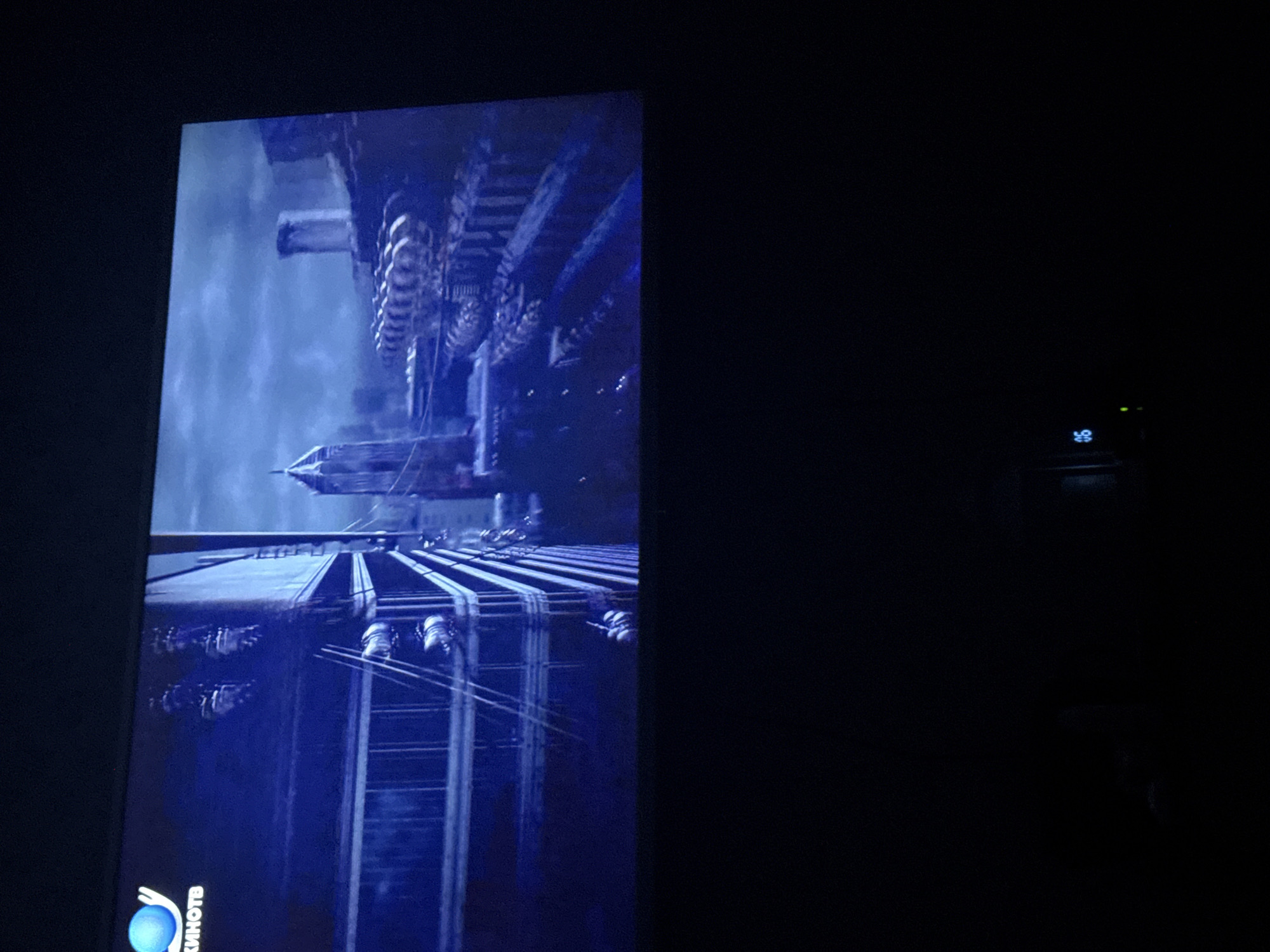 A monitor is displaying a scene from the film "Mortal Engines." The image shows a futuristic or steampunk-style city structure with detailed gears, buildings, and transport lines. The logo of Kinotb, a film distribution company, is visible in the bottom left corner. The environment is dark, with only the monitor illuminating the scene. The location, Mytishchi, Russia, is not visually apparent within the image. The scene is focused on the technological and architectural elements of the depicted city.