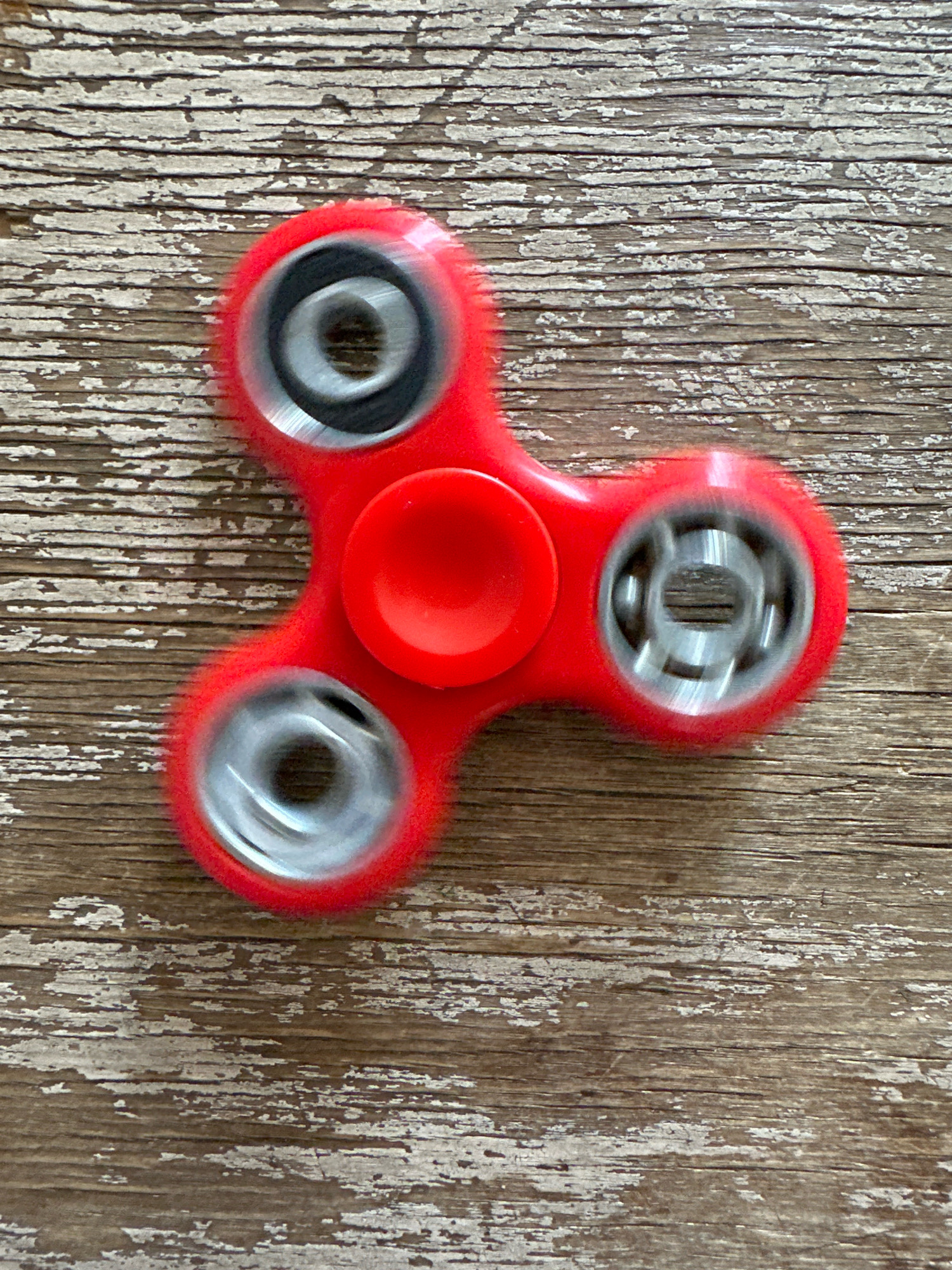 A red spinner toy is spinning on a weathered wooden surface. The spinner has three lobes, each with a bearing. The bearings are blurred due to the motion. The wooden surface has horizontal grain and patches of peeling white paint. The image is shot from directly above. There are no people or animals present. This scene does not have any specific identifiable markers indicating a location in Biysk, Russia.