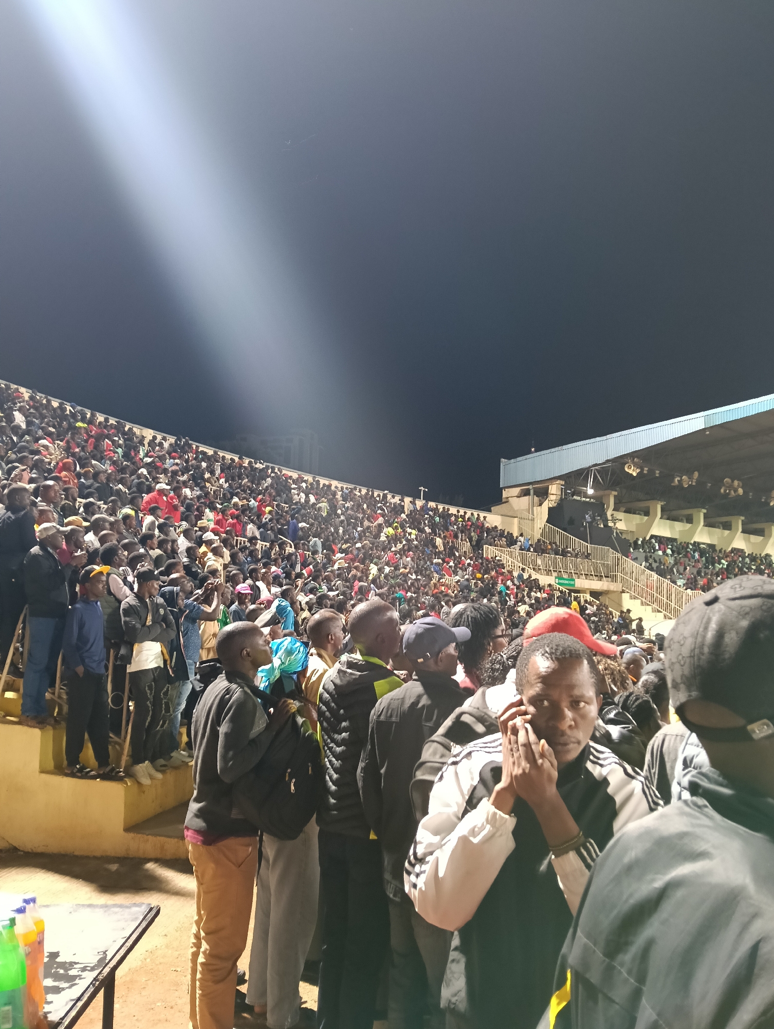 The image depicts a large crowd gathered in an outdoor stadium at night, with the location context provided as Nairobi, Kenya. The stadium's tiered concrete seating is densely populated with numerous individuals, primarily men, many of whom are looking upwards and towards a central area not visible in the frame.
A prominent, bright spotlight beam cuts diagonally across the dark night sky from the upper left, indicating that an event, such as a concert, sporting match, or public rally, is either in progress or about to commence. The sky is clear, suggesting fair weather.
In the immediate foreground, a man wearing a black and white track jacket is seen looking directly towards the viewer, holding a phone to his ear with both hands. His expression is focused. To his left, a man in a black jacket and light brown pants is visible, also observing the scene. On the far left, a table holds a few plastic bottles, possibly soft drinks.
The stadium architecture includes a light-colored structure with a blue roof section on the right side, along with visible staircases and railings. The scene is well-lit by artificial lights, highlighting the crowd and parts of the stadium against the dark expanse of the night sky. The large number of attendees suggests a significant public gathering. Faint, unreadable text on a distant green sign, possibly indicating an "Emergency Exit," is barely discernible within the stadium seating.