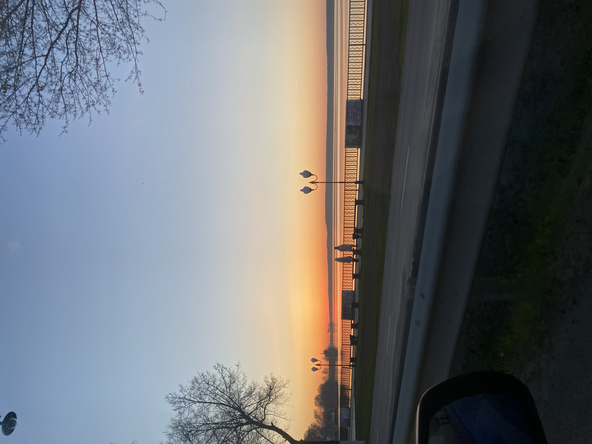 A photograph shows a street scene in Votkinsk, Russia, during sunset. The image is taken from a vehicle, as indicated by the visible side mirror and door frame. The sky transitions from blue to orange hues, reflecting on the water body to the right. Along the waterfront, there is a decorative metal fence and a row of lampposts. Silhouetted figures of people walking or standing are visible along the waterfront promenade. Bare trees frame the top and bottom of the image. A road runs alongside the waterfront.