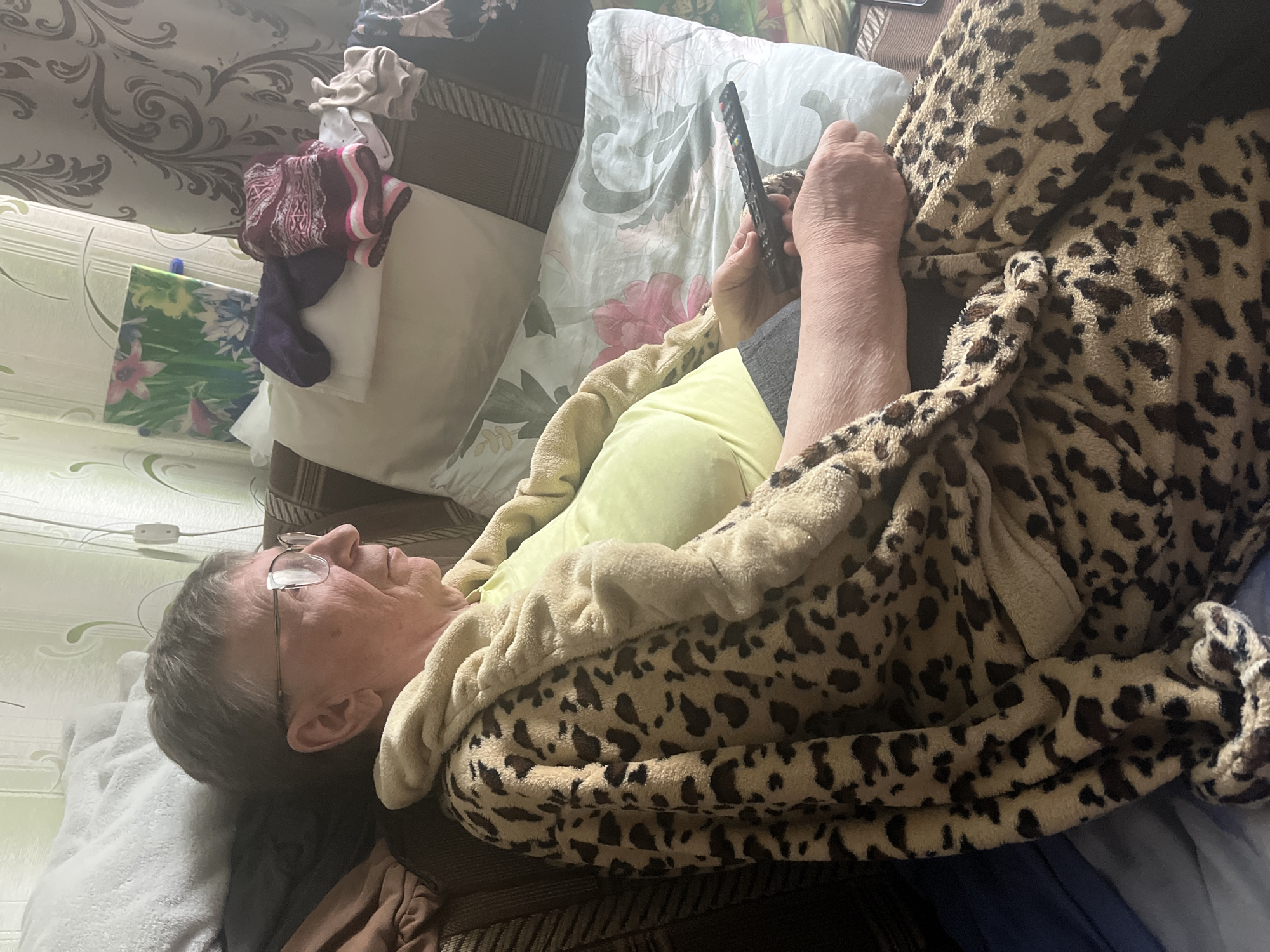 A person wearing a leopard-print robe and a lime green shirt sits on a couch, holding a TV remote. The person is also wearing glasses and has short, gray hair. The couch is covered with pillows and blankets. The scene appears to be an indoor setting, possibly a living room.