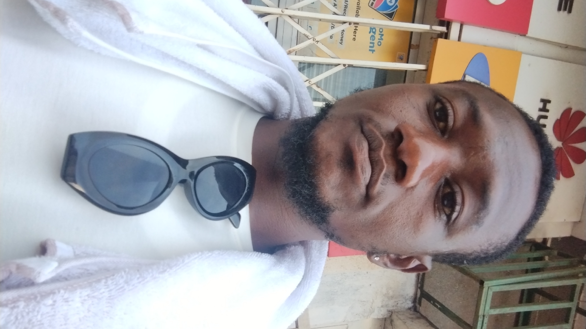 A close-up selfie of a Black male, appearing to be in his late 20s or early 30s, is presented. He has short black hair and a beard. He is wearing a white t-shirt and a white towel draped around his neck and shoulders. Black sunglasses are resting on his chest. In the background, there are signs with branding, including a prominent "HUAWEI" logo and what appears to be telecommunication company branding. The environment suggests an urban setting, possibly a commercial area, with a metal gate or bars visible. The overall impression is that the photo was taken outdoors, possibly in front of a shop or business establishment, within the city of Jos, Nigeria, given the context.