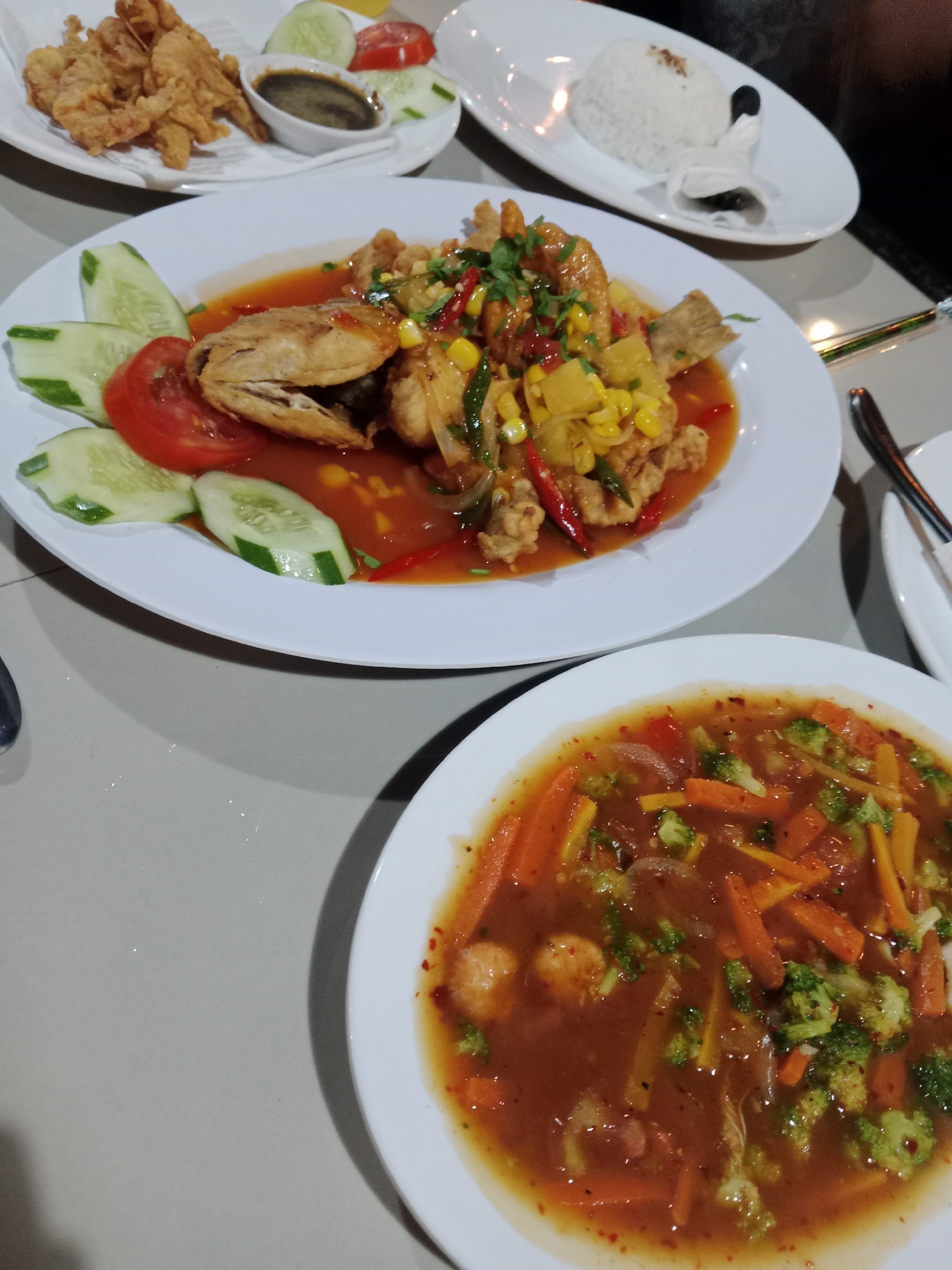 The image depicts a table in a restaurant setting, with several plates of food. In the foreground, there is a plate of what appears to be vegetable stew with visible carrots and broccoli in a reddish-brown sauce. Behind this is a plate with a fish dish, served with a tomato-based sauce, corn, and garnished with what seems to be sliced cucumber and tomato. Further back, there's a plate of fried food with a small bowl of sauce and additional cucumber and tomato slices. A plate of white rice is also visible. The table surface appears to be a light grey or off-white color. Cutlery is seen on the right side of the frame, partially obscured by the plates. The image lacks information about the location in the city of Unknown, Unknown, and contains no visible people or interactions.