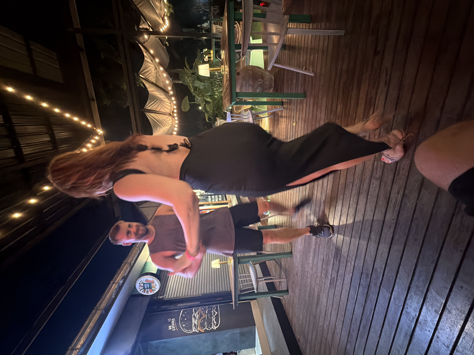 This media file captures a nighttime scene at what appears to be an outdoor dining or social venue in Baan Bang Rak, Thailand. Two people are visible: a woman in a long black dress and a man in a grey tank top and black shorts.
The woman, with long reddish-brown hair, is positioned in the foreground, her body angled towards the viewer. She is wearing high-heeled sandals. The man is in the mid-ground, facing the camera with a slight smile and beard. He is wearing athletic shoes.
The setting is a wooden deck with string lights illuminating the area. In the background, there are tables and chairs, some of which appear to be green, suggesting a casual outdoor restaurant or bar. Lush green plants are also present, adding to the tropical ambiance. The overall atmosphere seems relaxed and social.
The lighting suggests it is nighttime. There are no clear indications of the weather, but the outdoor setting implies it's likely pleasant.
Visible text includes "NO EXCUSES" written in a stylized font, likely part of signage for the establishment. There is also a circular logo with text that is difficult to decipher.