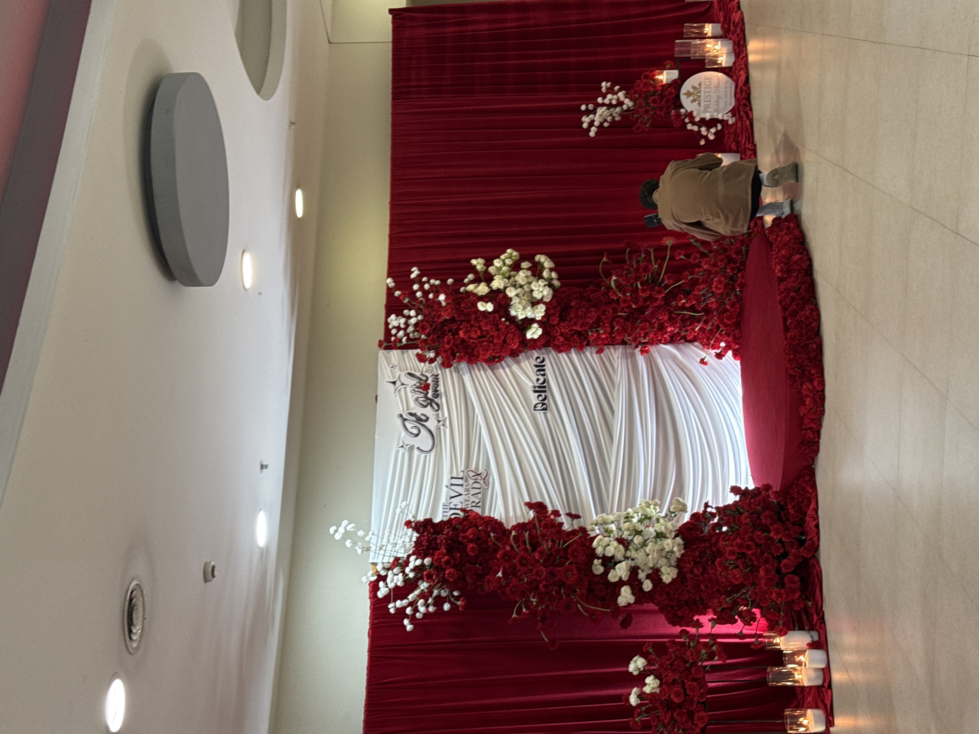 Here's a description of the image:
**Scene:** A photograph taken in a brightly lit indoor space, likely Yerevan, Armenia, shows a decorative setup for an event.
**Objects and Environment:**
* A large red curtain serves as a backdrop, draped with white fabric in the center.
* Floral arrangements of red and white flowers are prominent.
* Signage is present on the white fabric, reading "A Girl" and "The Devil Wears Prada".
* Candles in glass holders are placed on the ground near the curtain.
* The floor is a light tile or linoleum.
* The surrounding architecture includes white walls, recessed lighting, and decorative circular elements on the wall.
**People:**
* An individual wearing a tan jacket and dark pants is kneeling to the side. They appear to be interacting with the floral arrangements or the decoration.