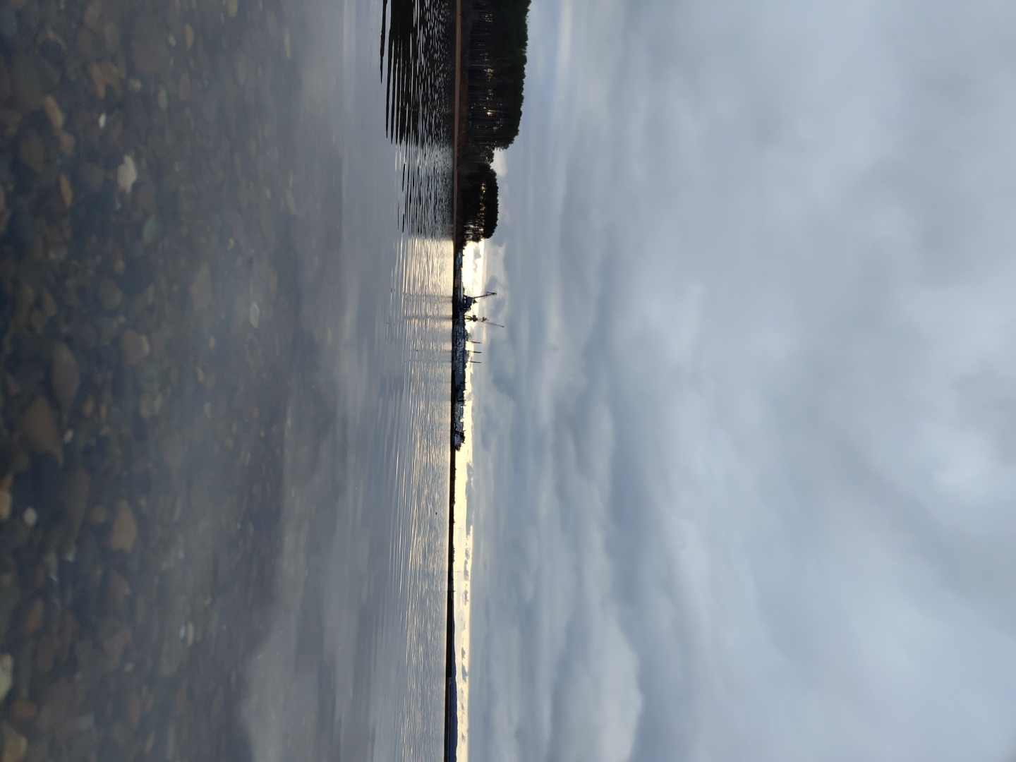 The image, correctly oriented, depicts a tranquil waterfront scene under an overcast sky. In the immediate foreground, clear, shallow water reveals a scattering of pebbles and small stones on the bottom. The surface of the water is calm, mirroring the sky and distant features.
Stretching across the midground is a broad expanse of water. On the distant right horizon, a dense line of dark evergreen trees marks the shoreline. Further along the horizon, a collection of structures, likely indicative of a port or industrial area, is visible, featuring several prominent cranes. A bright band of light reflects on the water in this distant section, suggesting the sun is low on the horizon, possibly rising or setting, and obscured by clouds or just out of frame.
The background is dominated by a vast, overcast sky filled with layers of soft grey clouds, creating a diffused and subdued lighting across the entire landscape. No people or specific activities are present in the scene. The overall impression is one of peaceful serenity and natural vastness.
The setting appears to be a large body of water, such as a lake or wide river, consistent with the location context of Chaikovsky, Russia, which lies on the Kama River and Votkinsk Reservoir. The time of day is likely early morning or late afternoon, characterized by the low sun's reflection. No text is visible in the image.