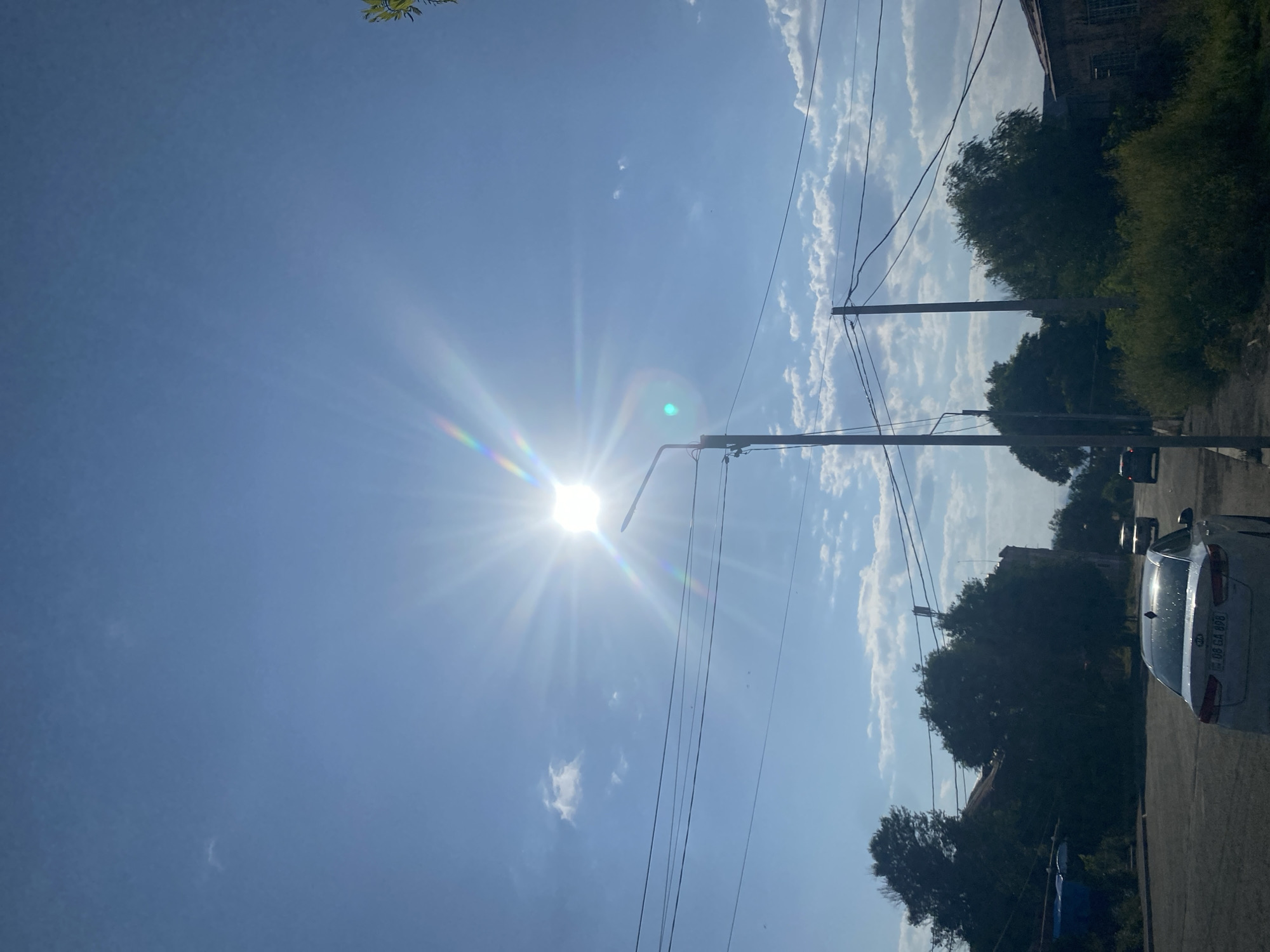 A low-angle, vertical shot captures a bright sun radiating light in a clear blue sky, with a few scattered clouds. The sun's rays create a rainbow effect, and a street light is visible in the foreground, positioned slightly to the right of center. The sky is filled with power lines running horizontally across the frame, supported by utility poles. Below the sky, a street scene unfolds with trees lining the road, and several cars are visible, including a white sedan in the foreground with a license plate reading "06 GA 898". Buildings are present on both sides of the street, some partially obscured by trees. The overall impression is of a sunny day in a suburban or semi-urban area.