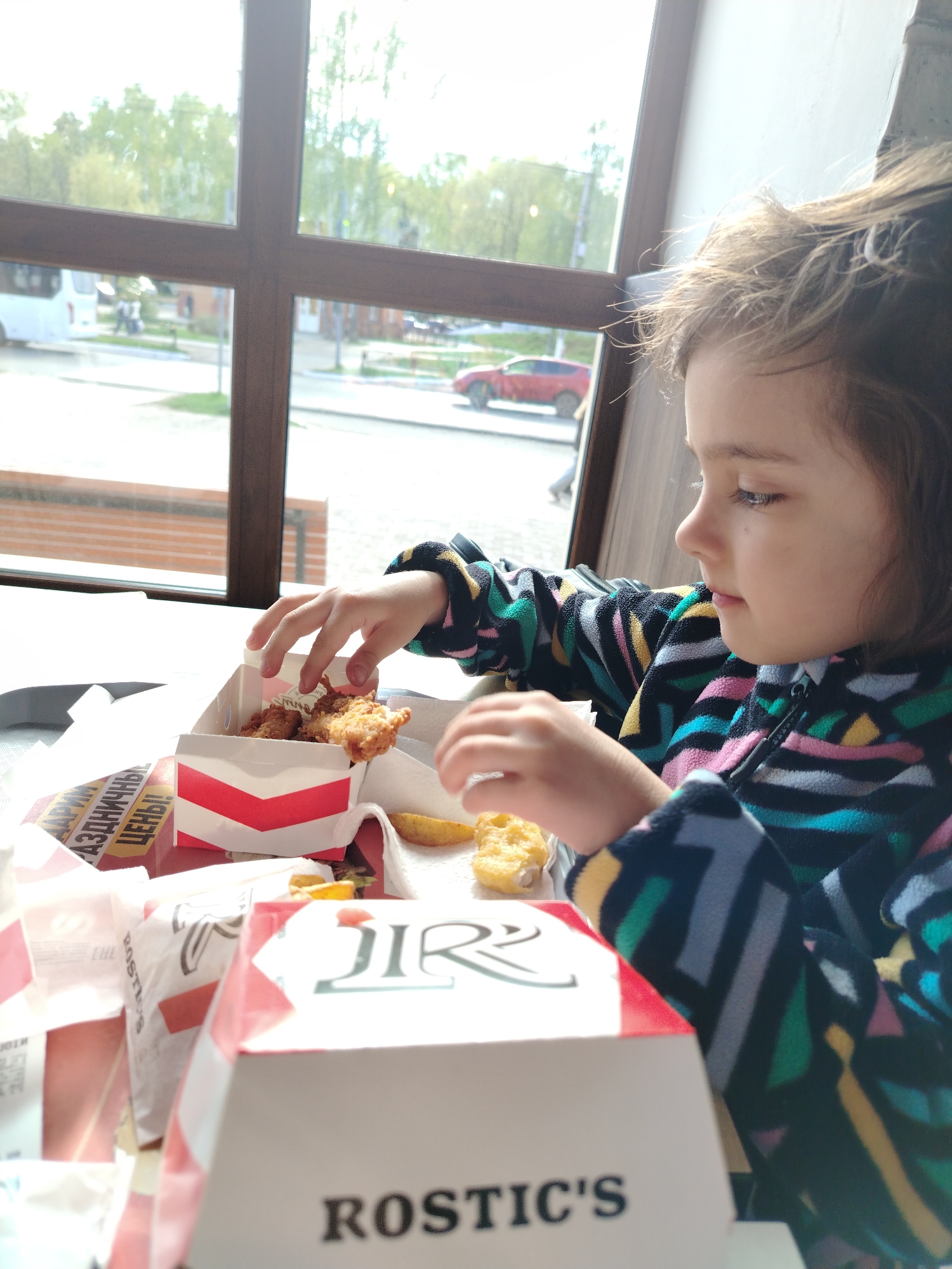 A young child is seated at a table inside a Rostic's restaurant, seemingly in Chaikovsky, Russia. The child is eating fried chicken from a Rostic's box. In the background, a window reveals a street scene with cars and buildings.
