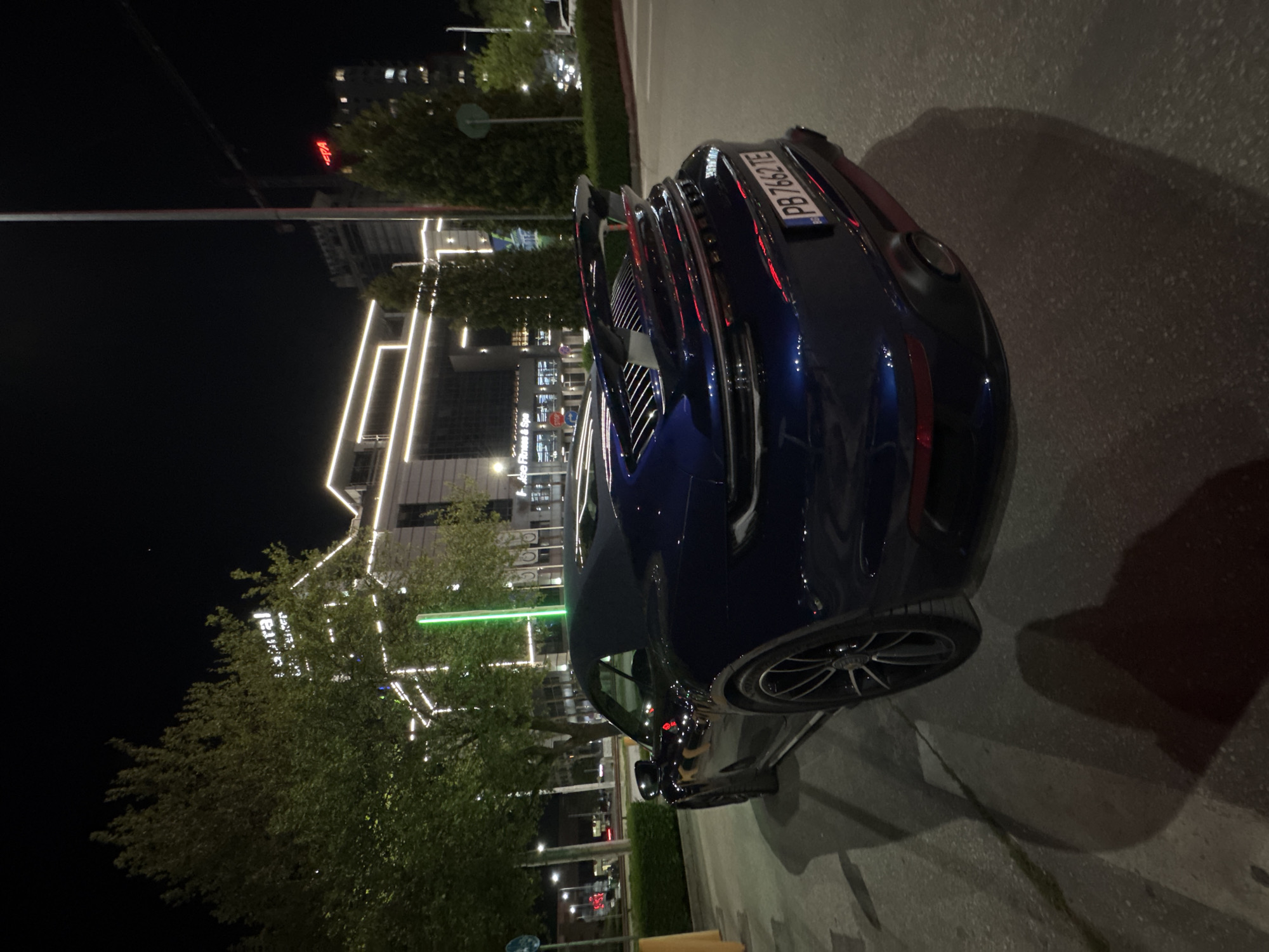 An outdoor nighttime photograph captures the rear three-quarter view of a dark blue, sleek sports car, likely a Porsche 911, parked on a paved surface. The car is positioned on the right side of the frame, angled towards the left, showcasing its distinctive rear spoiler, a full-width LED light bar, and a dark-colored wheel with silver accents on the right rear tire. The license plate, "PB 7687TE," is clearly visible on the rear of the vehicle, indicating its registration in Plovdiv, Bulgaria.
The background depicts a modern urban setting illuminated by various artificial lights. Tall buildings, some with distinctive vertical light strips, rise into the dark night sky. Lush green trees, also adorned with decorative white string lights, are interspersed between the buildings. A prominent green light pole or illuminated feature stands to the left of the car.
Visible text in the background includes "Ace Fitness & Spa" on a lower building and the word "Capital" written vertically on another taller structure. In the distant background, a small red illuminated sign is partially visible. The scene is calm, characteristic of a nighttime urban environment, with no discernible activity or people. The lighting from the buildings and streetlights creates reflections on the car's polished surface and casts shadows on the ground.