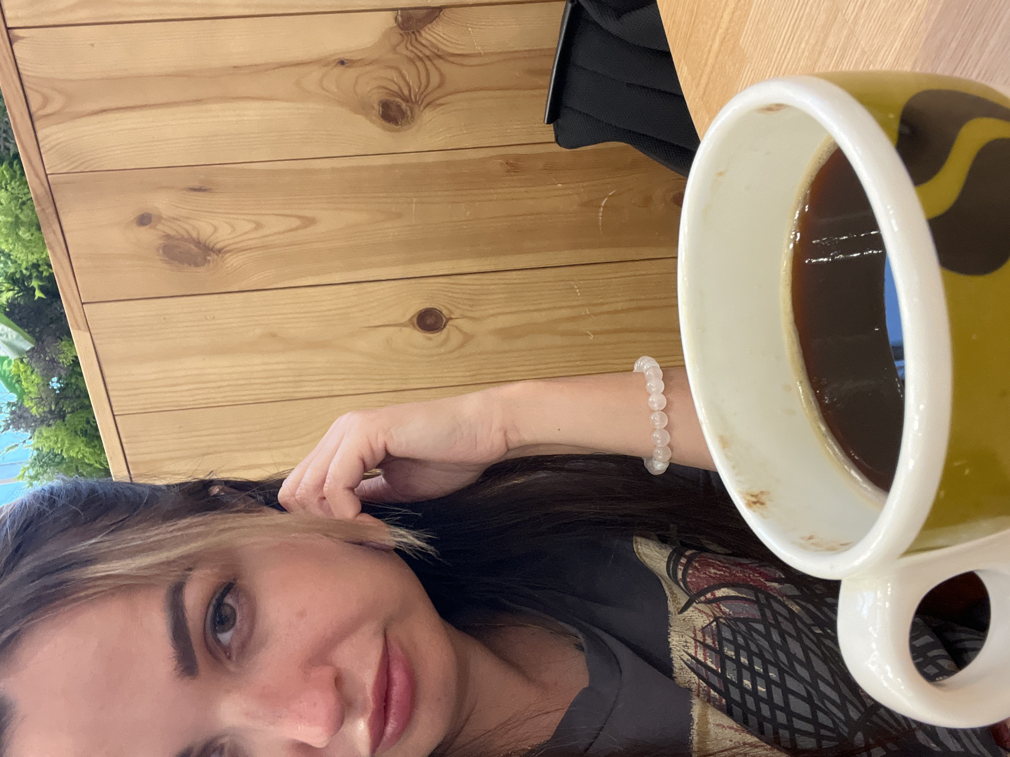 A close-up selfie shows a woman with long dark hair with blonde highlights sitting at a wooden table with a mug of coffee. She is wearing a dark t-shirt and a bracelet on her left wrist. The background is a wooden wall with some greenery at the top.