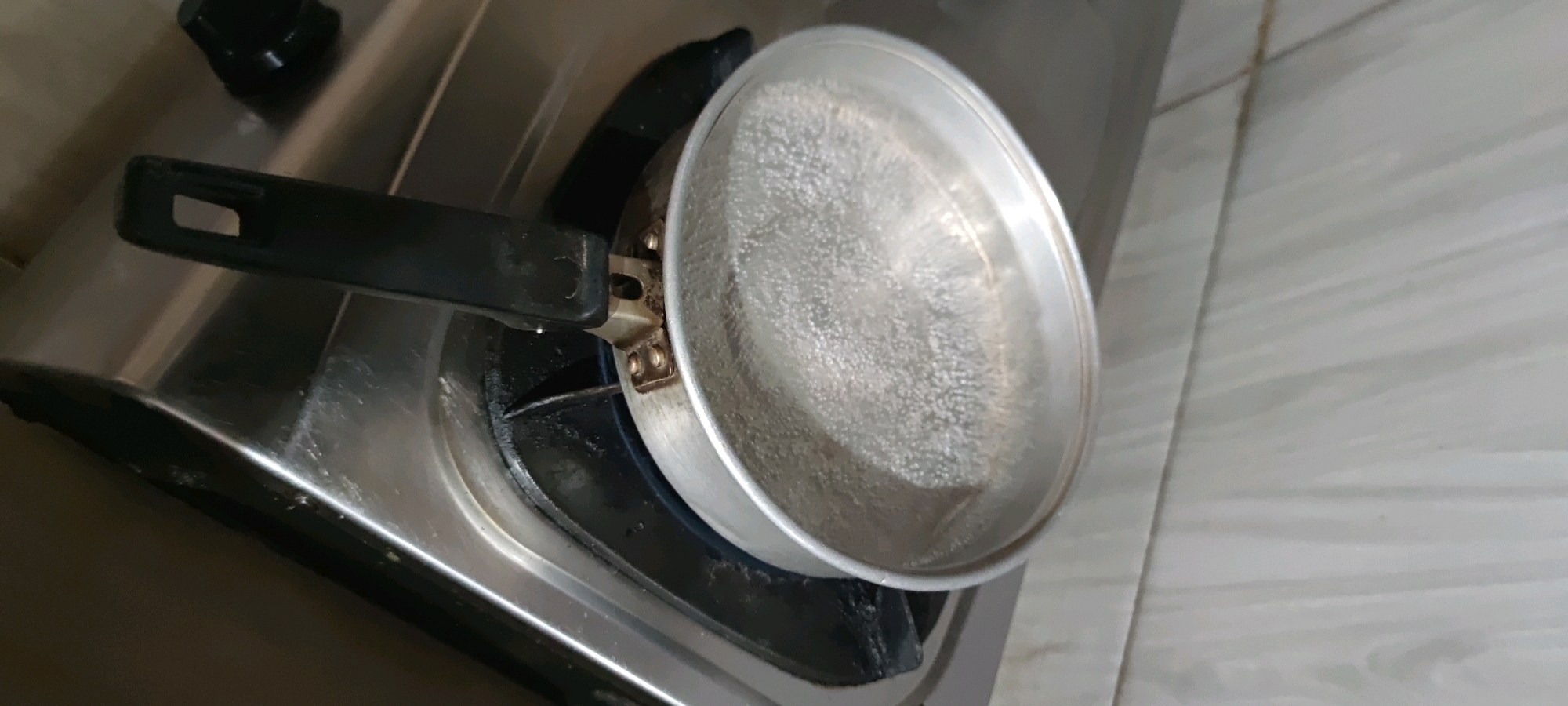 A close-up shot shows a pot of boiling water on a gas stove. The pot is made of aluminum and has a black handle. The stove is stainless steel and appears to be old and well-used, with some staining around the burner. The background consists of a tiled wall, which is light in color with a subtle pattern. The scene is set in Gambarsari, Indonesia.