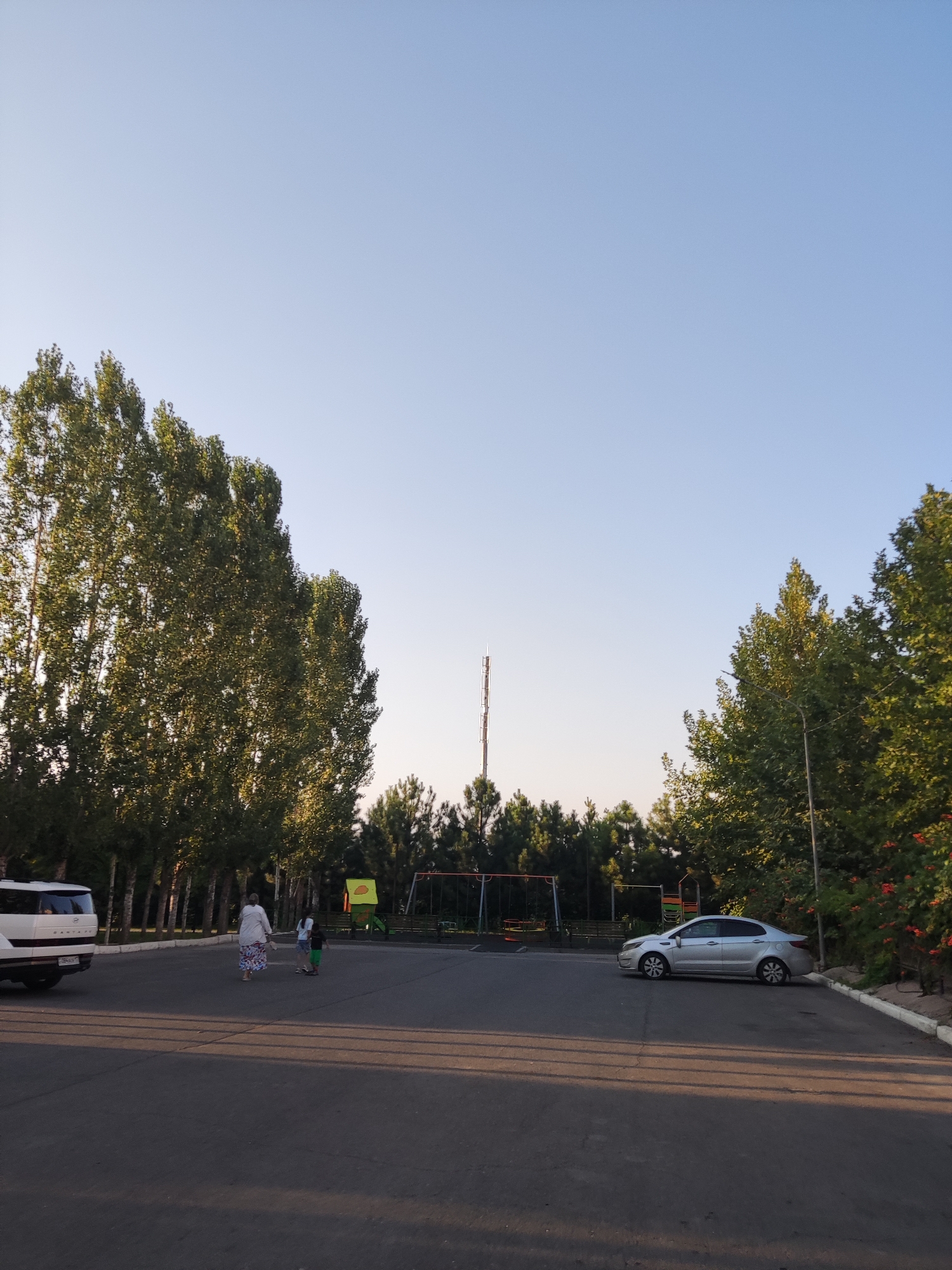 The image depicts an outdoor scene with a clear blue sky. In the foreground, a paved area, possibly a parking lot, is visible, with shadows cast from trees. On the left side, a white SUV is parked. Further into the scene, two individuals, an adult and a child, are walking. In the background, a playground area with swings and a climbing structure is present, surrounded by a line of tall trees. Beyond the trees, a telecommunications tower stands tall, and on the right side, a silver sedan is parked. The environment appears to be a park or recreational area within a city, with the presence of urban infrastructure and greenery. The specific location is identified as the city of Shymkent, Kazakhstan.
