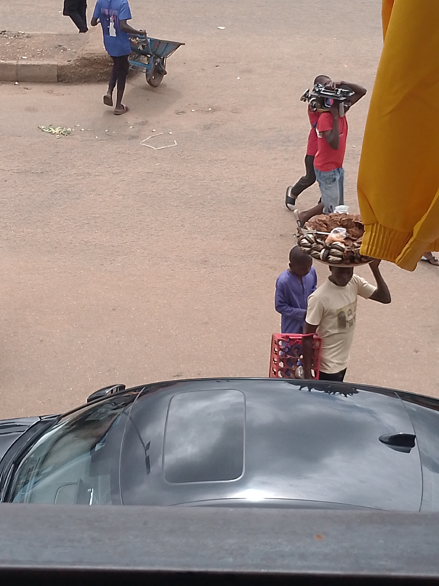 In Bukuru, Nigeria, a scene unfolds on a dusty, paved street. In the foreground, the roof of a black car with a sunroof is visible. Further up the street, two young men are walking. One, in a light-colored t-shirt, carries a large, round tray piled high with food items on his head. Beside him, a younger boy in a blue tunic walks with his head down. In front of them is a red plastic crate.
In the background, another young man in a red t-shirt and denim shorts is walking, carrying a complex piece of electronic equipment on his shoulders, possibly a camera rig. Behind him, another person in a blue shirt pushes a wheelbarrow. The street is unpaved in places, with scattered debris and a few green leaves on the ground. A large expanse of yellow fabric, possibly a flag or tarp, hangs into the frame from the right side. The overall environment suggests a busy, possibly informal market or commercial area.