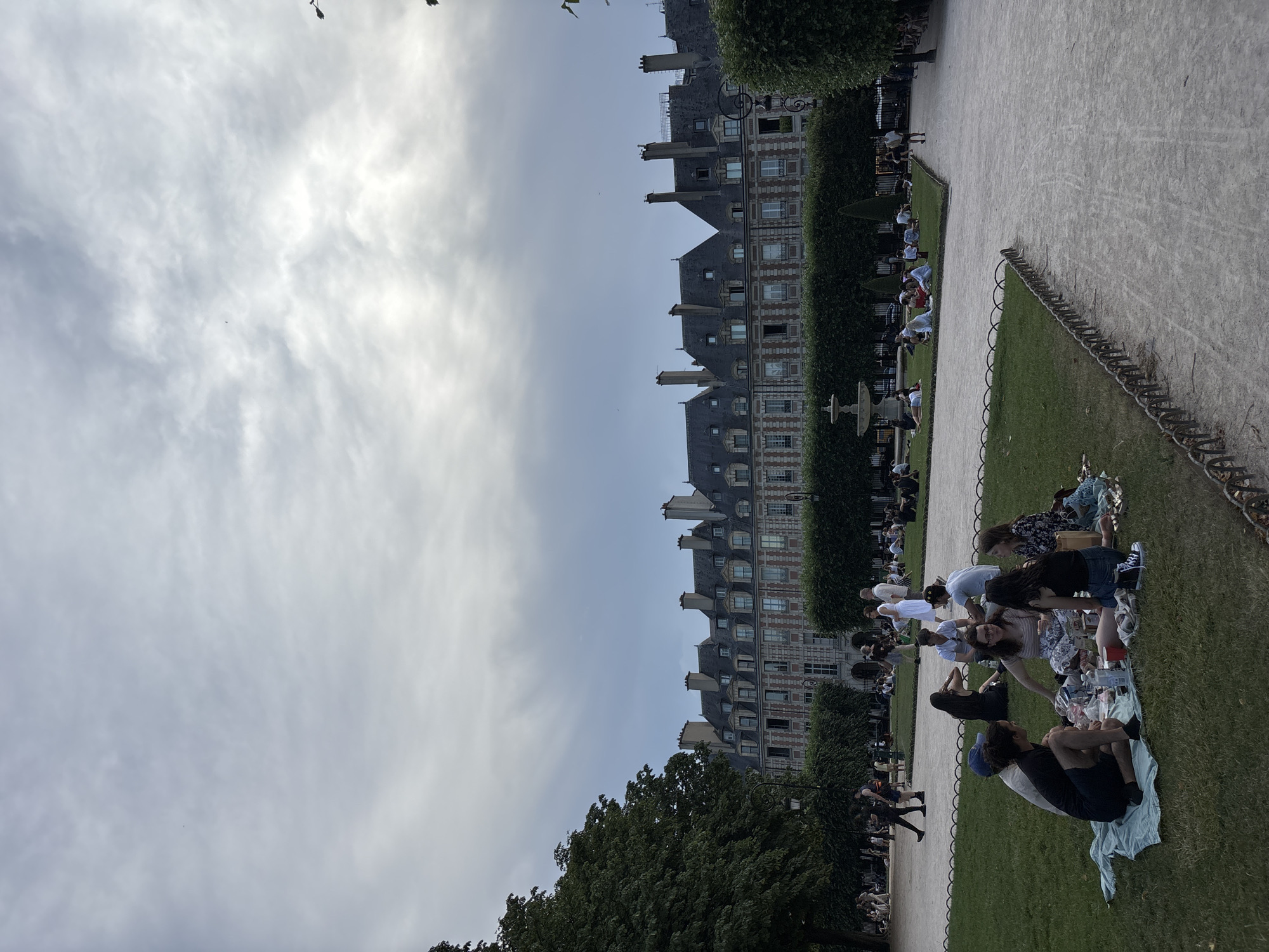 The image depicts a public park in Paris, France, likely the Place des Vosges, with people enjoying a picnic and the general ambiance of the square. In the foreground, a group of seven people are seated on a light blue blanket on a grassy area, engaged in conversation and eating. Some are holding drinks, and food items are spread across the blanket. To the left of this group, a tall, dark green tree provides shade. In the middle ground, several other individuals and small groups are scattered across the lawn, some also picnicking, others walking or standing. A fountain is visible in the distance, surrounded by manicured hedges. Behind the hedges, a row of ornate, historical buildings with mansard roofs and dormer windows line the square. The sky is overcast with a mix of grey and lighter clouds, suggesting either early morning, late afternoon, or an impending weather change. The overall scene conveys a relaxed and leisurely atmosphere.
