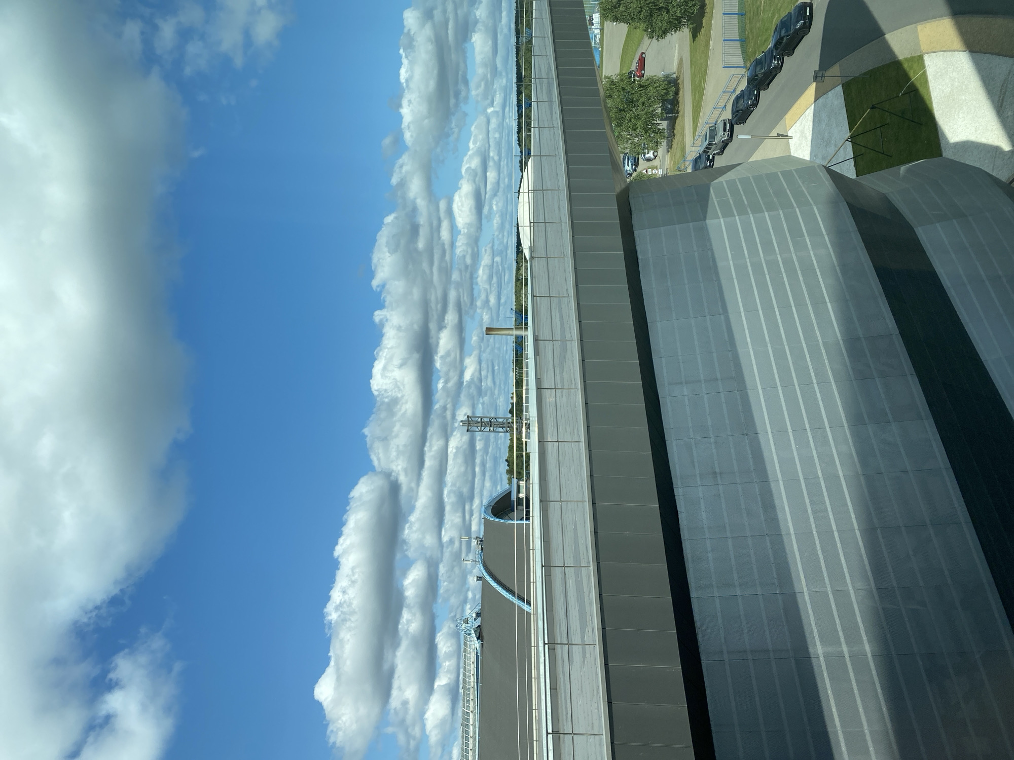 This is a photograph of the National Aero Space Center of Belarus, located in Minsk, Belarus, on a sunny day with scattered clouds. The foreground features a large, modern building with a grey, curved facade. Above this building, a walkway with railings stretches across the frame, connecting to another structure. Beyond the walkway, a large, dark-roofed building with arched blue accents is visible, along with a tall industrial tower in the distance. The sky is a vibrant blue, filled with fluffy white and grey clouds. In the lower right corner, several cars are parked on a street adjacent to a green lawn and trees.