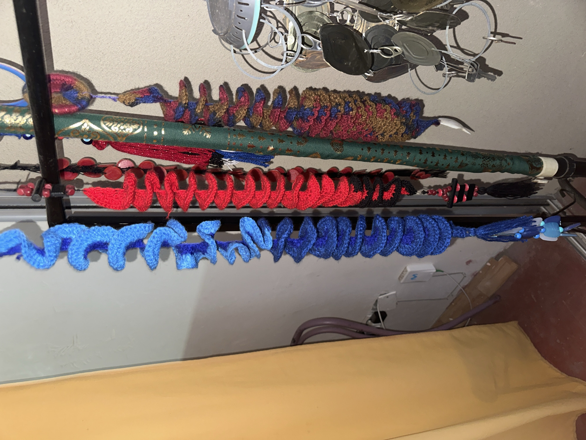The image presents a cluttered scene with multiple objects suspended from a metal rod against a light-colored wall. A multi-colored crocheted item, a green and gold patterned tube, and a red and black garland with beaded embellishments are visible. A bright blue crocheted garland is suspended from the lowest rod. Above these are suspended metal utensils or decorative items. Below, there is a golden or beige surface, possibly a bed or a piece of furniture. The location and city of Arica, Chile is not definitively indicated within the visual content.