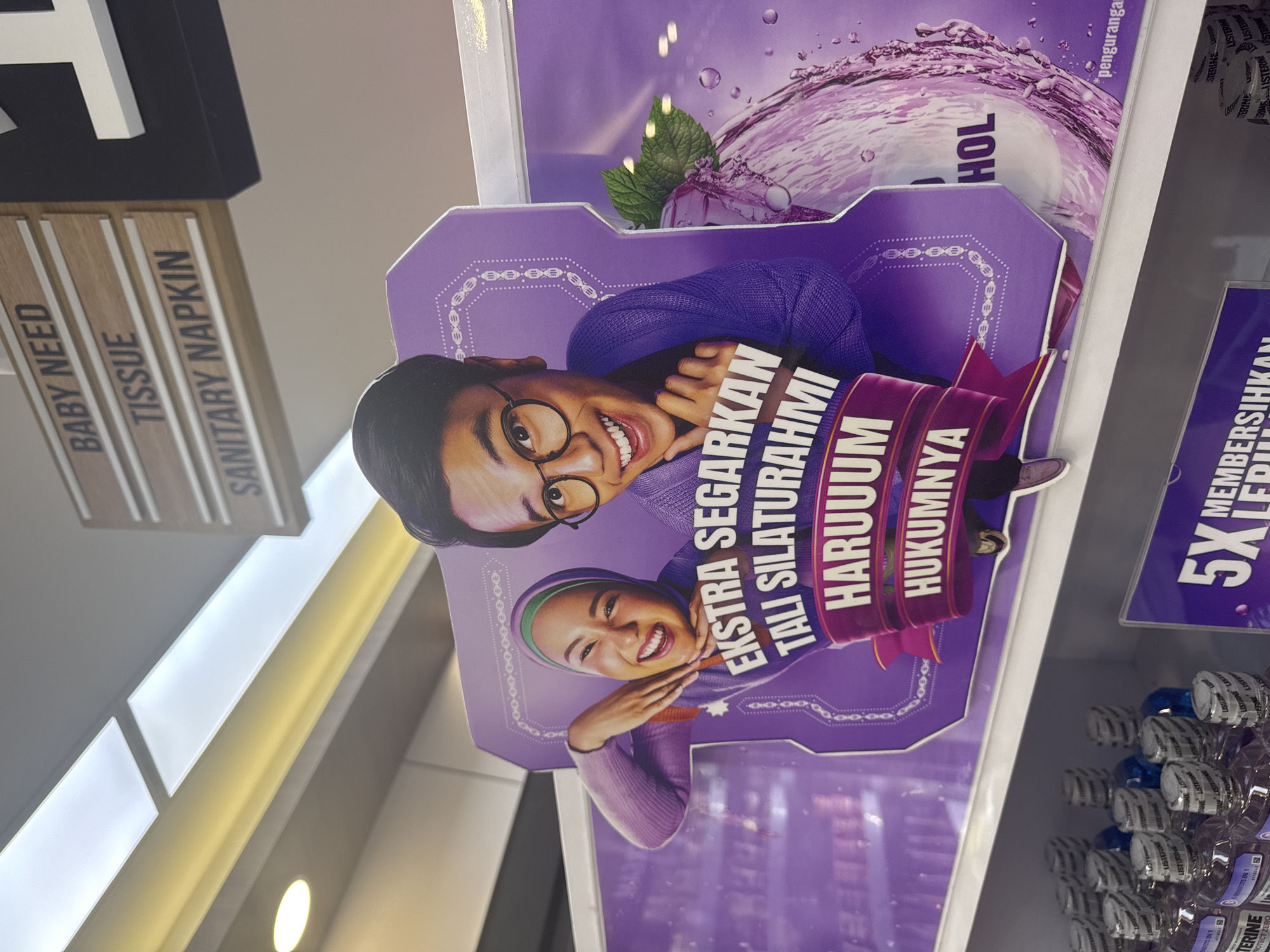 This rotated image captures an indoor retail setting, likely an aisle in a supermarket or pharmacy in Kuta Selatan, Indonesia. The primary focus is a purple, stand-alone promotional display featuring two smiling individuals – a man with glasses and a woman wearing a hijab. Both appear happy and friendly, conveying positive emotions.
The display features a prominent slogan in Indonesian: "EKSTRA SEGARKAN TALI SILATURAHMI HARUUUM HUKUMNYA." This translates to "Extra refresh ties of kinship/social bonds, fragrant/pleasant is its ruling/law," likely a marketing phrase encouraging fresh breath for social interactions, common around holidays in Indonesia. Behind this cutout, a larger purple graphic depicts a splash of liquid with mint leaves, and partially visible text "HOL" (likely part of "ALCOHOL" or "NON ALCOHOL") and "penguranga" (reduction).
To the left, light wooden signs are visible, labeled vertically with "BABY NEED," "TISSUE," and "SANITARY NAPKIN," indicating product categories in the store. To the right, shelves are stocked with numerous bottles of Listerine mouthwash, identifiable by the repeated "LISTERINE" text, including variations like "BENEFITS IN 1" and "PROTECT ZERO." Some bottles contain blue liquid, others appear to have a purple hue. Another partial sign on the right mentions "MEMBERSIHKAN" (cleaning) and "5X," possibly referring to a cleaning benefit. The scene highlights a consumer product display within a busy retail environment.