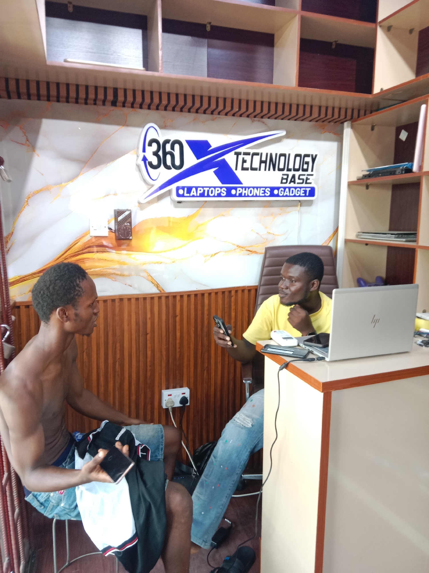 The image shows the interior of a small technology shop, possibly in Onitsha, Nigeria, given the context. Two men are present. One man, shirtless, sits on a metal chair with clothing in his lap and is holding a mobile phone in his hand. The other man, wearing a yellow t-shirt and blue jeans, is seated at a desk in front of a laptop. He is looking at the shirtless man and holding a cell phone in his hand. The desk contains various items, including a laptop, a remote, and charging cables. The shop's signage on the wall reads "360 Technology Base" with the words "Laptops, Phones, Gadget" underneath. Shelving units are visible in the background. An electrical outlet is on the wall behind the shirtless man.
