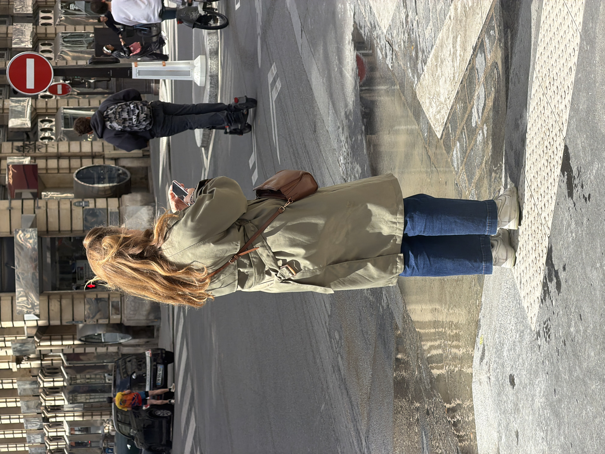 The image presents a tilted, eye-level perspective of a bustling street scene in Paris during daylight hours.
In the immediate foreground, a person with long, wavy light brown hair is visible from the back. They are wearing an olive green trench coat, blue jeans, and white sneakers, and a brown leather shoulder bag hangs across their body. This individual is holding and appearing to interact with a smartphone.
Further back on the sidewalk, a second person wearing a dark hoodie and a black and white patterned backpack walks away from the viewer. On the street, a cyclist in a white shirt and dark pants can be seen. In the background, near a parked black car with the license plate "CN-554-GU", two other individuals are present; one is notably wearing a colorful shirt featuring yellow, red, and blue.
The street features a grey asphalt road with white markings and a paved sidewalk, part of which includes tactile warning strips for the visually impaired. Buildings with modern, reflective, or metallic facades line the left side of the street. A prominent red circular "no entry" sign (French "sens interdit") with a white horizontal bar is clearly visible, with a smaller similar sign beneath it. The bright lighting indicates daytime, likely sunny, with visible shadows. The overall scene depicts typical urban activity with people walking, cycling, and engaging with mobile devices.