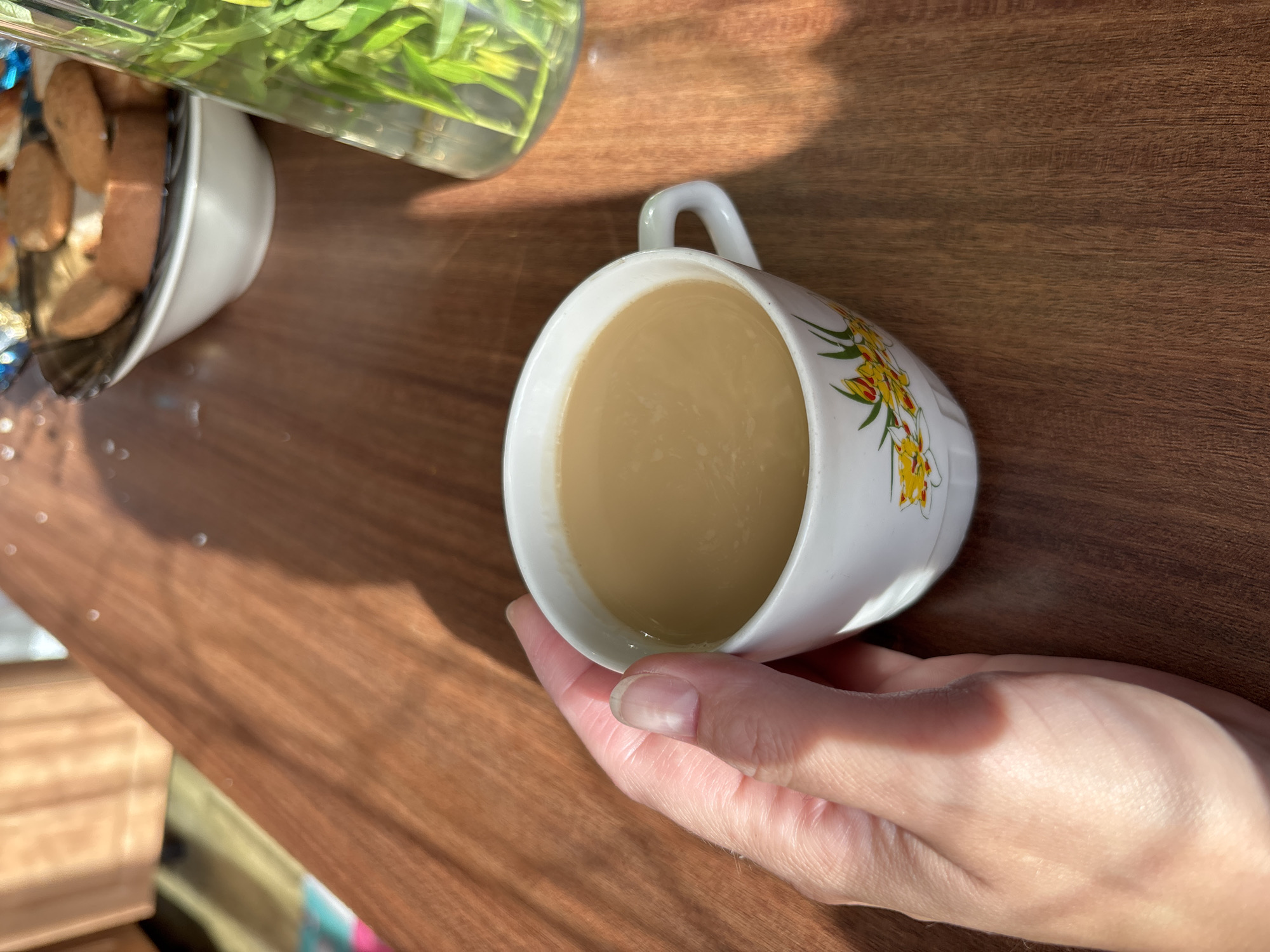 An over-the-shoulder shot captures a hand holding a white ceramic mug filled with light brown liquid, likely coffee or tea, with a floral design on its exterior. The mug sits on a wooden surface, with a bowl of cookies and a glass vase filled with greenery in the background. Sunlight casts shadows across the scene, creating a warm, dappled effect. The image appears to be taken indoors, possibly in a home or cafe.
