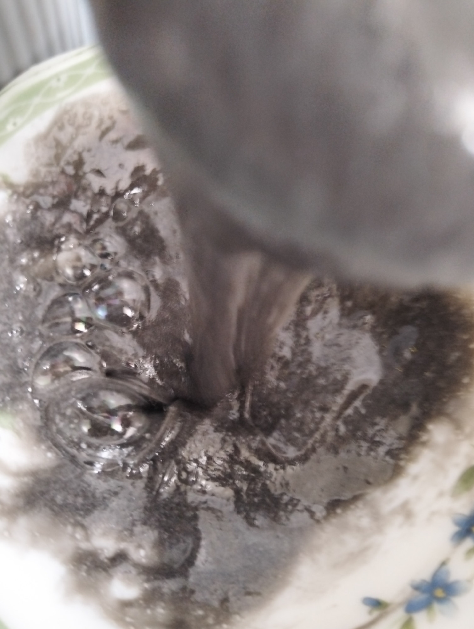 A close-up, slightly out-of-focus view, possibly taken with a mobile phone, shows a dark, viscous liquid with numerous air bubbles being poured into a white ceramic bowl. The bowl has a floral pattern with blue flowers and green leaves along its rim. Part of the object pouring the liquid is visible at the top of the frame and appears to be a dull, gray material. The liquid itself is dark gray or black and has a thick, almost sludge-like consistency. The bubbles suggest some degree of agitation or mixing. The image doesn't offer information that would allow locating it in Dhaka, Bangladesh.