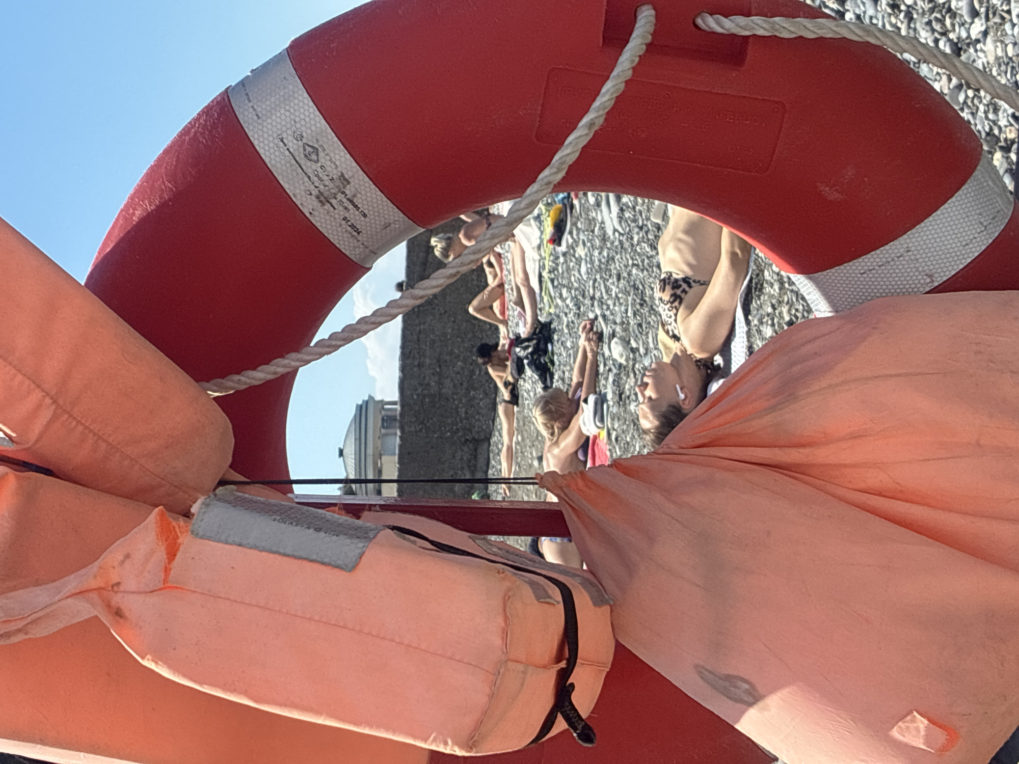 This image captures a sunny day at a beach, likely in Sochi, Russia, with a prominent red life preserver in the foreground. The perspective is from below, looking up towards a clear blue sky and the life preserver, which is looped by a thick white rope. Several people are visible in the background, seemingly enjoying the beach. Some are lying down, possibly sunbathing, while others appear to be sitting or standing. One person in the background is wearing a bikini with a leopard print pattern.
In the immediate foreground, partially obscuring the scene, are orange life vests. The life preserver itself has reflective silver bands with visible text and markings, including what appears to be a certification or manufacturer's label. The beach appears to be pebbly. The overall atmosphere suggests a relaxed, recreational setting during daylight hours, likely during warmer weather given the attire of the people. The sunlight is bright, casting shadows and highlighting the textures of the life vests and the life preserver. The background shows a hint of architecture, suggesting the beach is near an urban or developed area. The image conveys a sense of summer leisure and safety consciousness due to the presence of life-saving equipment.