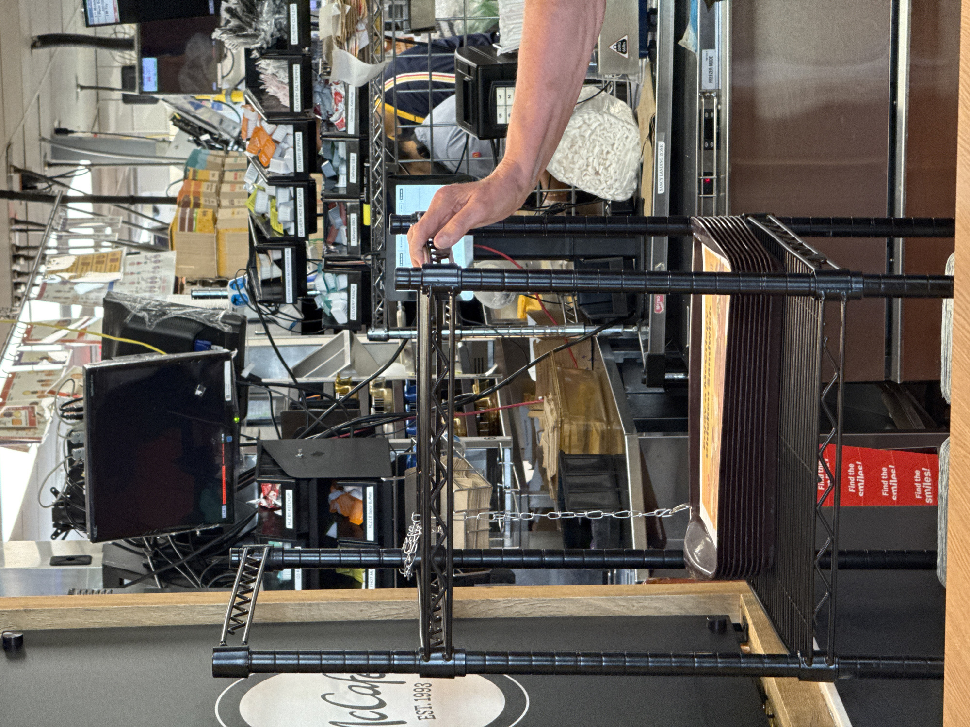 An eye-level shot captures a busy service or back-of-house area, likely in a fast-food restaurant or café such as a McDonald's, indicated by the partial "McCafé EST. 1993" logo visible on a counter. The setting is cluttered with numerous screens, wire shelving, and various operational components, consistent with a commercial kitchen in Columbus, United States of America.
A person's right arm and hand are prominent in the center, reaching towards and interacting with a small digital screen or device, possibly operating an order management system or point-of-sale terminal.
The space features black wire shelving units, some filled with plastic containers organized for "DELIVERY" sauces, including "KETCHUP," "Spicy Buff" (likely Buffalo), "HOT MUSTARD," "BUFFALO," "RANCH," "BBQ," and "S&S" (Sweet & Sour). Multiple computer monitors, some displaying interfaces, are stacked and surrounded by a dense network of cables. Other visible objects include a numerical keypad, a bag of white, possibly packaging material, and a metal chain with a scoop hanging from it.
Signs within the scene include "NANCY LANDING ZONE ONE" and "FREEZER MODE." Red placards prominently display the repeated message "Find the smiles!". The overall impression is that of an active and operational commercial food service environment.