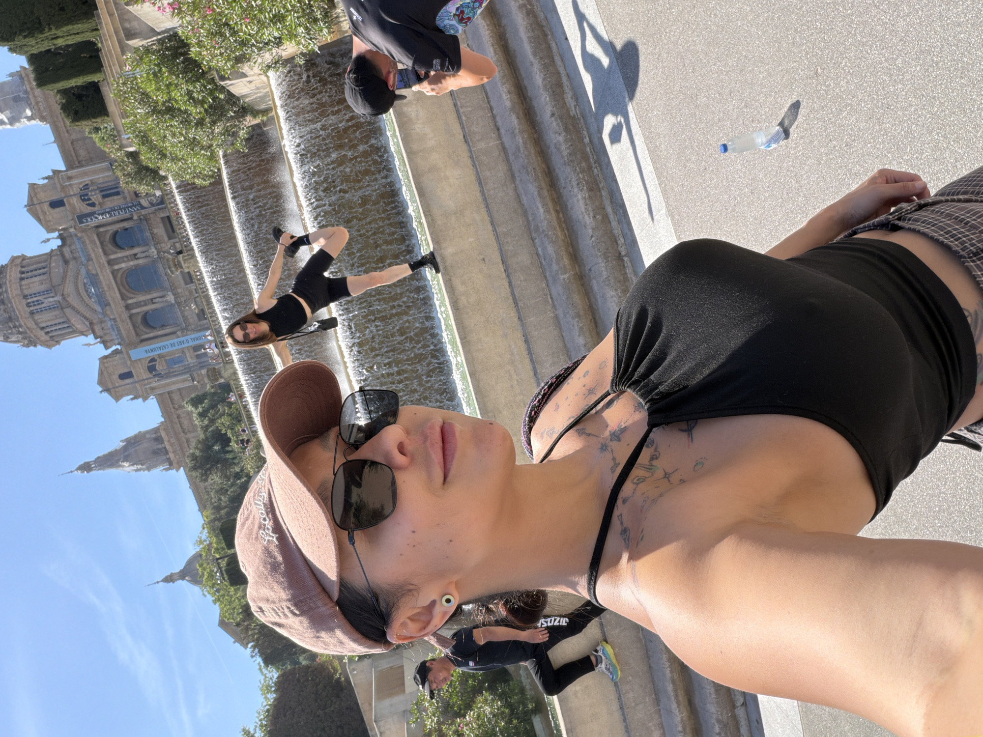The image shows a woman taking a selfie at the Magic Fountain of Montjuïc in Barcelona, Spain. The fountain, a tiered structure with cascading water, is visible in the background. The woman is wearing a hat, sunglasses, and a black top. Other people are in the background, including one posing for a photo in the fountain and another taking a picture. A water bottle is on the ground.