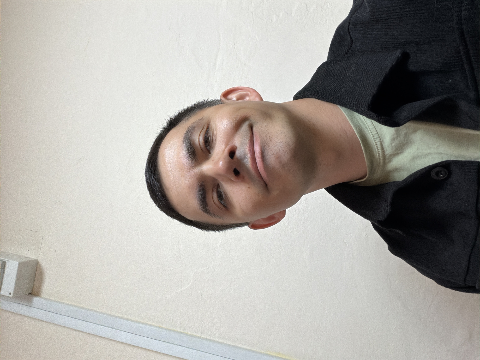 A close-up, tilted, upward-facing selfie captures a person with short, dark hair and light skin. They are wearing a light green t-shirt and a black corduroy jacket. The background is a plain, light beige wall with a white conduit running horizontally along the bottom. There is no discernible information to identify this location as Izhevsk, Russia, or any other specific city.