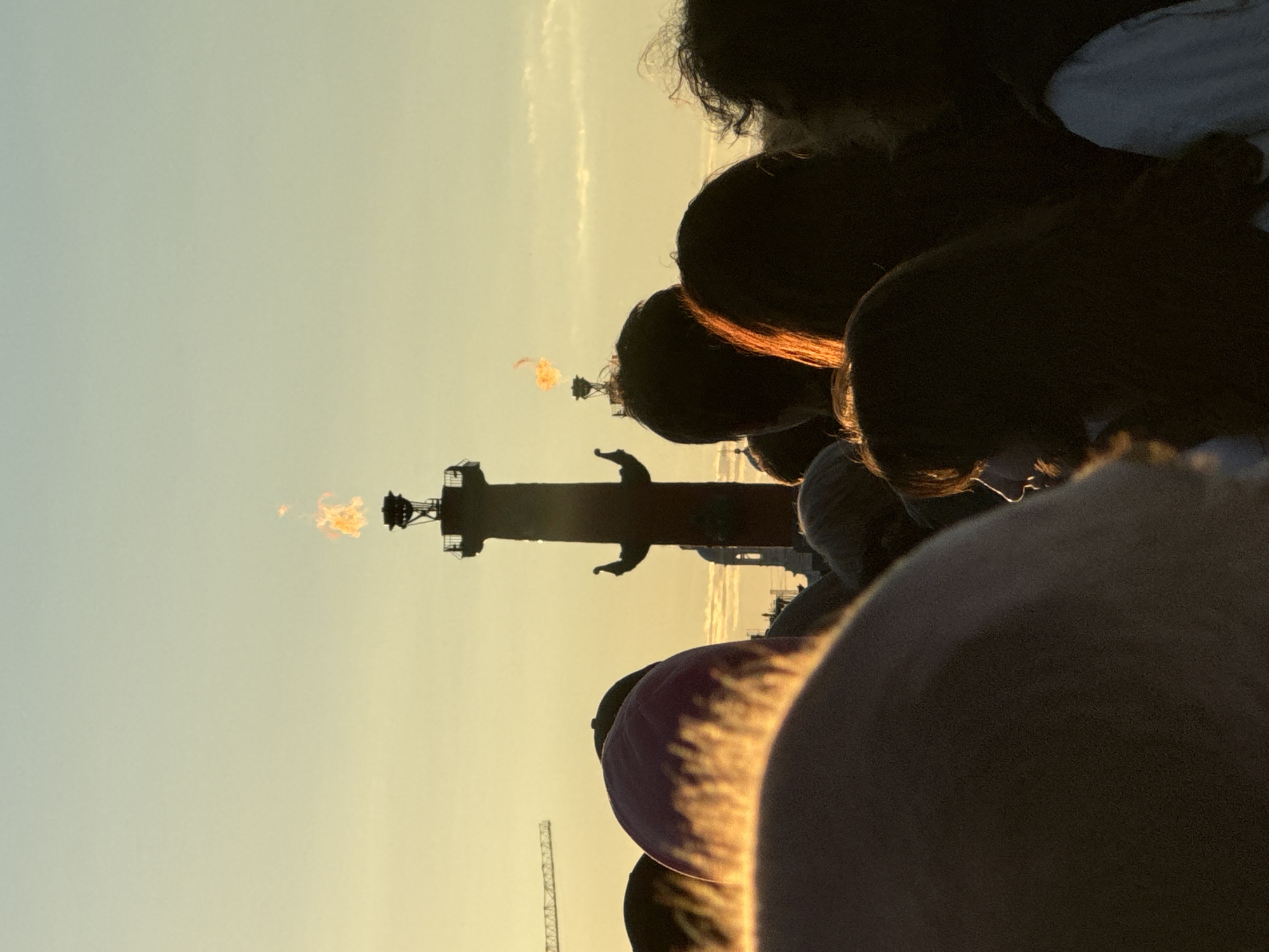The image depicts a crowd of people silhouetted against a warm, golden sunset sky in Saint Petersburg, Russia. In the background, a tall, slender red column topped with a flaming beacon stands prominently. The flame flickers with vibrant orange and yellow hues. To the left of the column, a construction crane is visible against the hazy sky. The foreground is dominated by the backs and sides of several individuals, their faces turned away from the camera, suggesting they are all looking towards the beacon or the sunset. One person in the middle ground is turned slightly to the left, and their profile is visible, with the golden light highlighting their hair and facial features. The overall atmosphere is one of gathering and observation, possibly for a public event or simply to admire the sunset over the city.