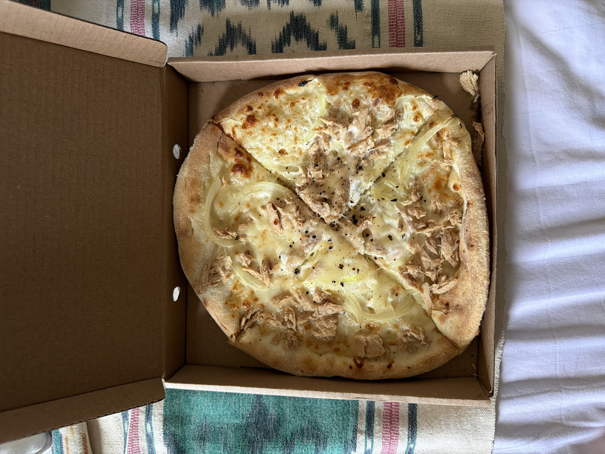 The image shows a pizza in a cardboard pizza box. The pizza is cut into four slices. Toppings appear to include cheese, tuna, and onions. Some slices have black pepper. The pizza box is open, with the lid partially visible. The background consists of a patterned fabric. The image was taken in North Kuta, Indonesia.