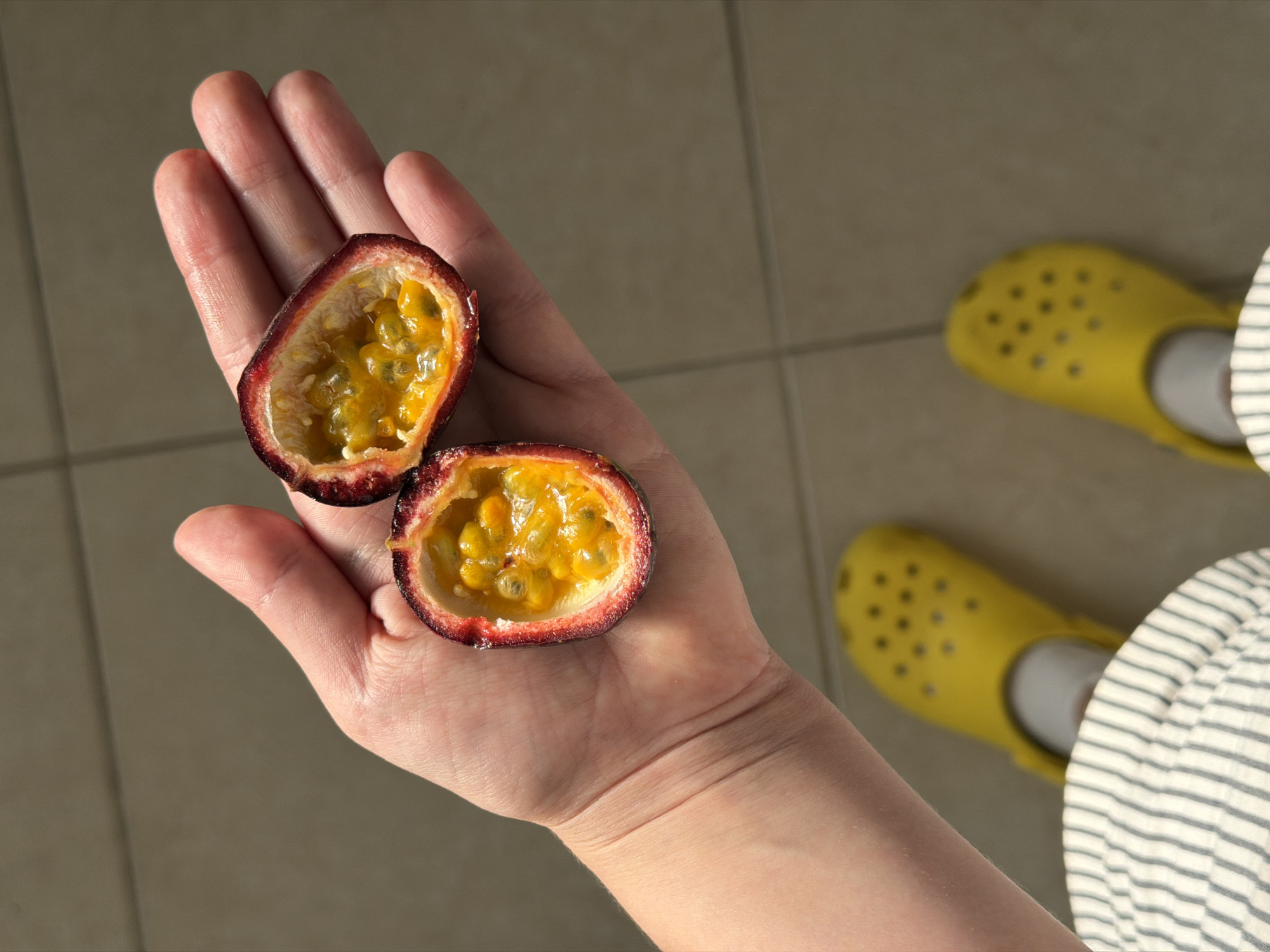 A person's hand holds two halves of a passion fruit. The fruit's interior is pulpy with seeds, and the rind is a deep red. The person is wearing a striped garment and bright yellow Crocs on their feet. The location appears to be indoors, possibly a home, evidenced by the tile flooring. There's no visible information that confirms the location as the city of Tel-Aviv, Israel.