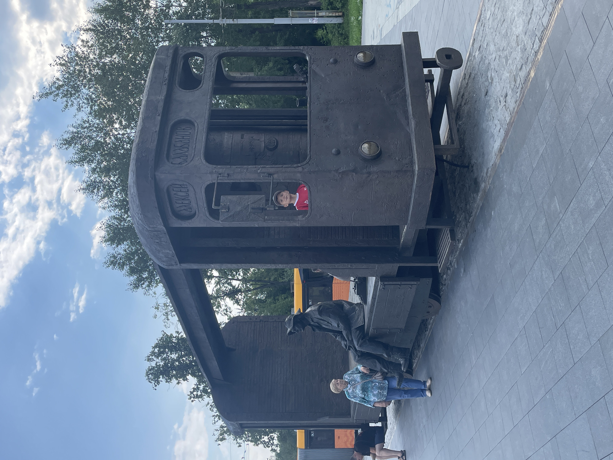 The image depicts an outdoor public space, likely a street or park, in Yekaterinburg, Russia, featuring a monument to Aleksei Balabanov. The monument is a large, dark-colored sculpture resembling a stylized train car. One section of the sculpture is open, revealing a seated figure of a man in a hat, seemingly a representation of Balabanov. To the left of this figure, an older woman in a blue patterned shirt and jeans stands, looking towards the sculpture. A young boy in a red shirt is visible inside the cabin of the train sculpture, looking out. In the background, trees line the horizon, and a modern tram or train with yellow and orange accents is visible on a track to the left of the monument. The ground is paved with large, grey square tiles in the foreground, transitioning to cobblestones near the monument. The sky above is blue with scattered white clouds.