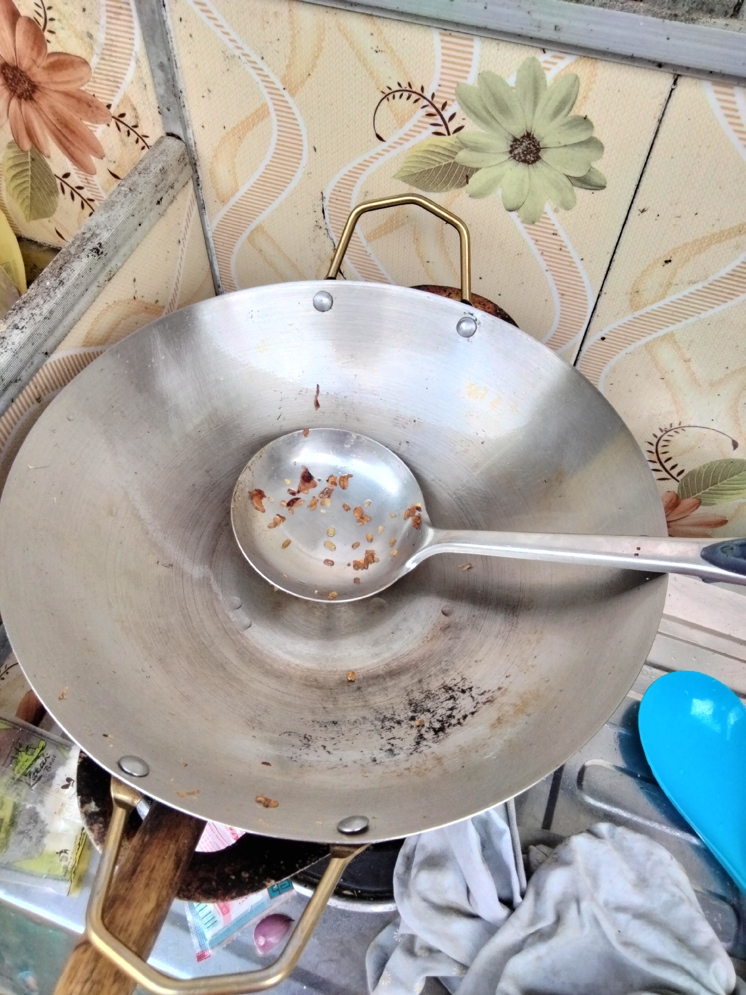 A wide shot, taken from above, shows a used wok with a ladle resting inside on a stovetop in Cariu, Indonesia. The wok is made of brushed metal and contains remnants of fried food. The ladle is also metal and has a blue handle. A blue plastic spoon is visible to the right of the wok. The background is a tiled wall with a floral pattern. The scene suggests a kitchen environment that may be outdoors or in a simple dwelling.