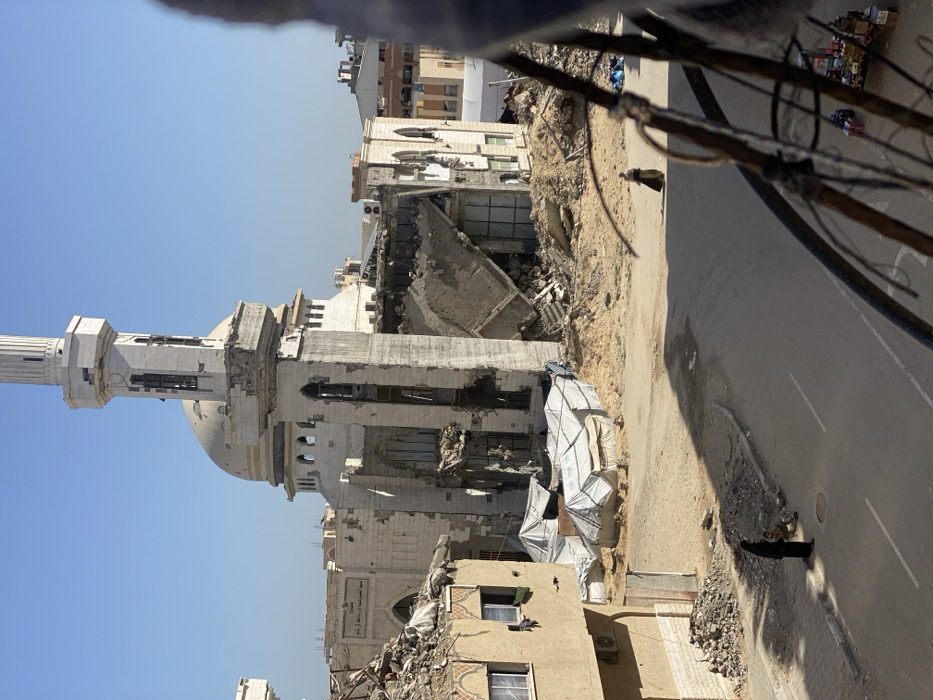 The scene depicts a street in Khan Yunis, Palestinian Territory, under a clear blue sky. The prominent structure in the mid-ground is the severely damaged Al-Farouq Mosque, identifiable by its distinct minaret and dome. The minaret exhibits significant structural damage to its upper sections. Large portions of the mosque's main building, including its facade and interior areas, are collapsed into extensive piles of concrete and rebar rubble.
To the left, a multi-story residential building shows broken windows and visible damage, with clothing hanging from a window. At its base, additional rubble is present. Between this foreground building and the mosque, a sandy area contains several improvised shelters constructed from white tarpaulins. To the right of the mosque, another building exhibits catastrophic collapse, with its internal structure protruding outwards.
A paved road runs horizontally across the lower mid-ground. On the left side of the road, a person in dark attire stands beside a small child seated on the ground near a pile of debris. Further right on the road, another person in dark clothing walks away from the viewer. The image is captured from an elevated position, with wires and structural elements of the viewpoint building partially visible in the immediate foreground on the right.