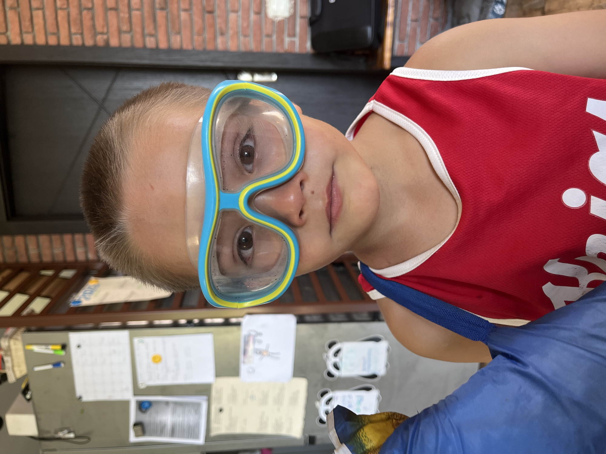 A young child wearing a red athletic tank top and blue and yellow swim goggles is pictured indoors. The child is holding a blue object, possibly a bag or a sling, which obscures part of their body. In the background, a grey refrigerator with various papers and drawings attached is visible, along with a brick wall and a dark door. The scene appears to be a domestic setting, potentially a kitchen or hallway. The overall impression is of a candid, informal photograph. This image was taken in the city of Ureki, Georgia.