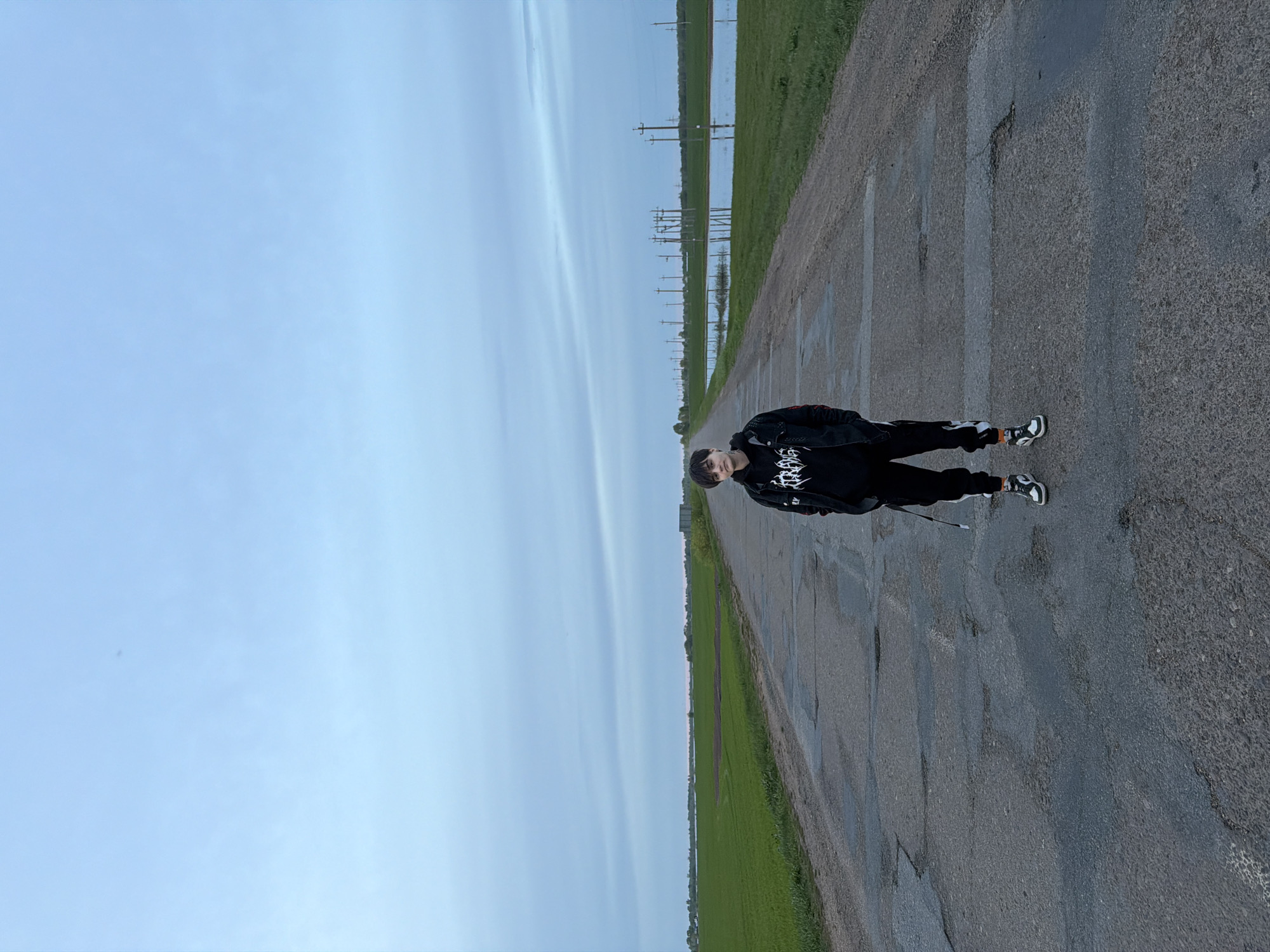 Azyakul', Russia: A person stands on an asphalt road. To the right of the road is green grass. To the left of the road is a field and a blue sky. Power lines can be seen in the distance.