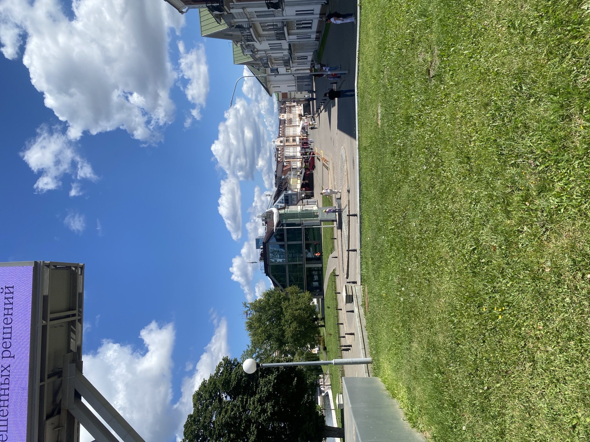 The image captures a sunny day in a city square, likely in Minsk, Belarus, given the presence of a billboard with Cyrillic text. In the foreground, a well-maintained green lawn slopes upwards, with some dandelions scattered throughout. A grey concrete structure lines the left edge of the frame, possibly part of a modern building or an urban design element.
Beyond the lawn, a paved area forms the main square. A tall, grey lamppost with a spherical white light fixture stands prominently in the mid-ground. Several small black bollards are arranged in a line along the edge of a raised flower bed or planter.
Further into the square, a modern building with a glass facade and dark framing is visible on the left. To the right of this building, a street lined with multi-story buildings stretches into the distance. These buildings exhibit a mix of architectural styles, with some featuring classical elements like balconies and ornate trim, while others appear more contemporary. The street is populated by a few pedestrians walking in various directions. A red car is parked on the side of the street.
The sky is a vibrant blue, adorned with fluffy white cumulus clouds, indicating fair weather. The overall impression is one of an active urban public space on a pleasant day.
