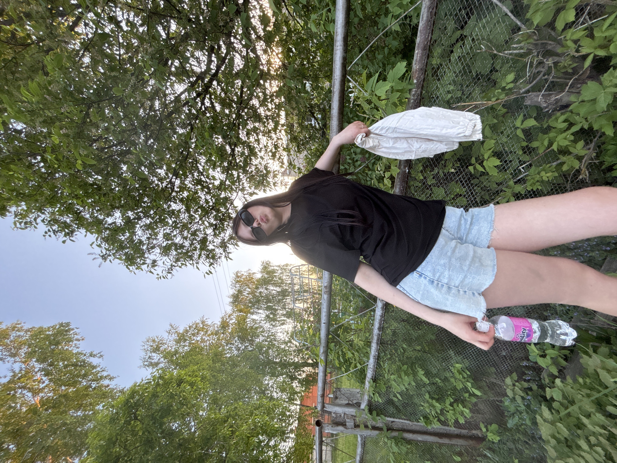 A young woman wearing sunglasses, a black t-shirt, and light denim shorts is seen standing near a metal fence. She is holding a white garment in her left hand and a plastic water bottle in her right. The backdrop features green foliage and trees, while a glimpse of the sky is visible above the trees. The location is in the city of Фоки, Russia.