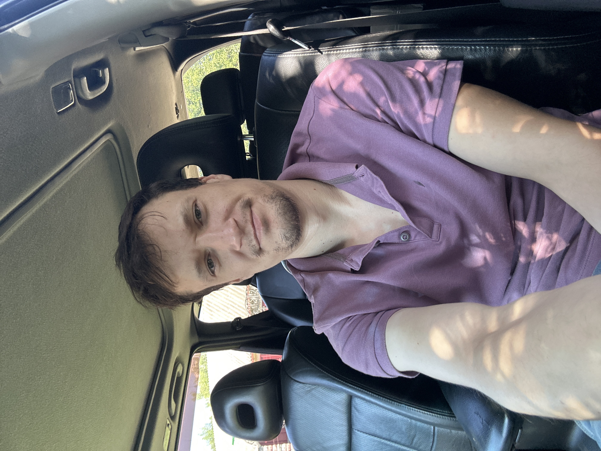 A young adult male with short brown hair and a light beard is seated in the driver's seat of a car. He is wearing a purple polo shirt and looking directly at the camera with a slight smile. The interior of the car is visible, including black leather seats, a sunroof, and the dashboard. Outside the car, trees and buildings can be seen, suggesting an outdoor location, possibly in Shatava, Ukraine, given the context. The image appears to be a selfie taken during the daytime, with sunlight casting shadows on the man's face and arm.