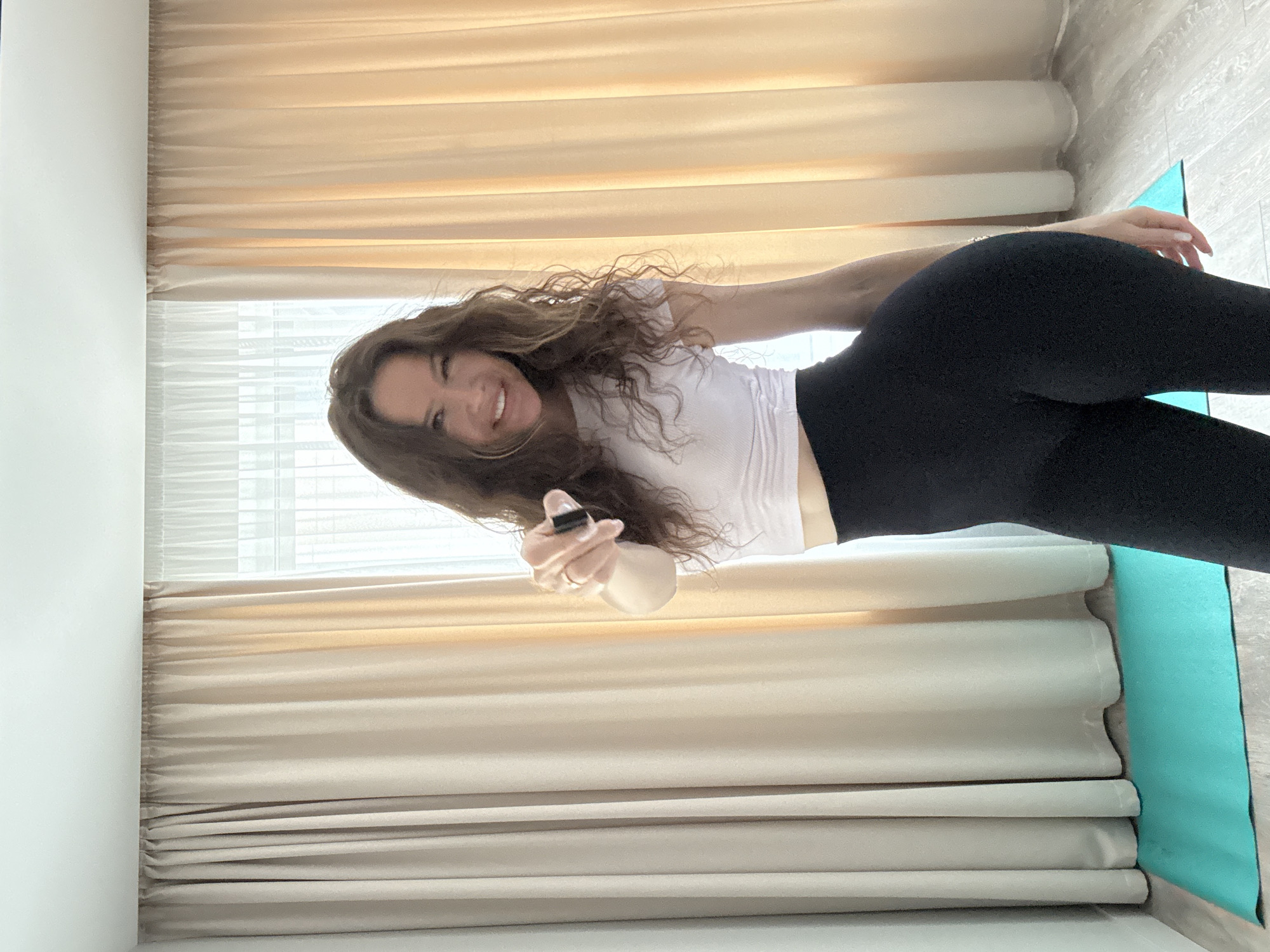 The image shows a woman indoors, standing in front of curtains. She is wearing a white crop top and black leggings. Her hair is long and wavy. She is smiling and pointing her finger towards the camera. There is a turquoise yoga mat on the floor to her left. The room appears to be a bedroom or living room, with light-colored walls and wooden flooring. The overall lighting suggests it is daytime. There is no indication of the location being Moscow, Russia.