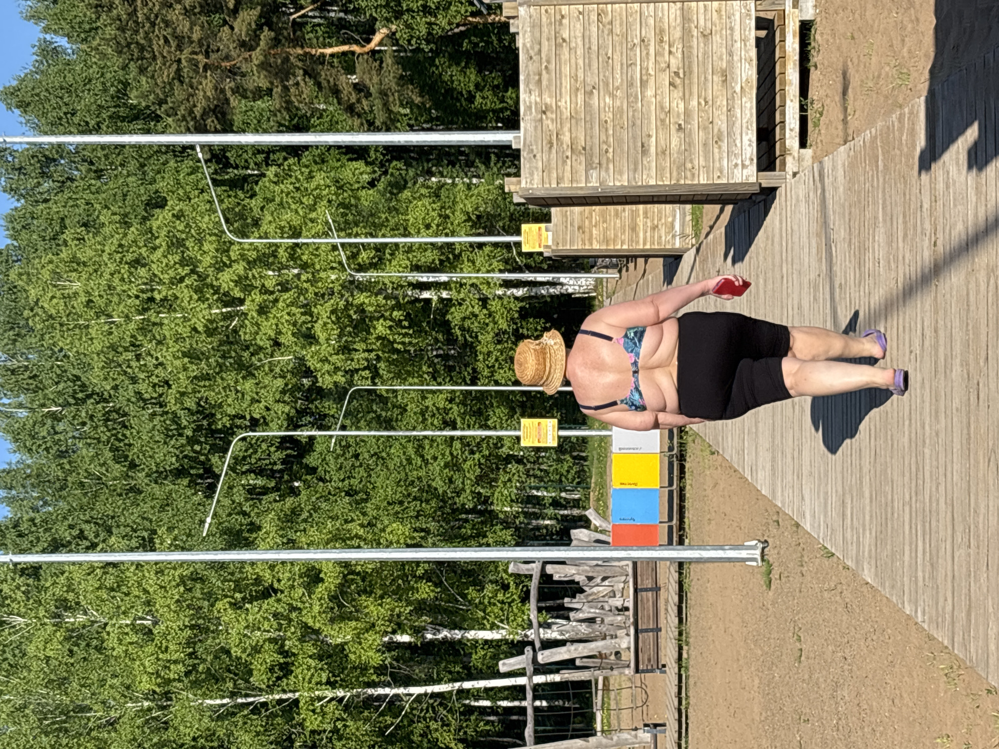 A woman wearing a straw hat, a bikini top, and black shorts walks along a wooden boardwalk. She holds a red object in her right hand. To the left, a series of metal posts and what appear to be informational signs stand amidst green trees. A wooden structure is visible in the background. This scene is located in the city of Chaikovsky, Russia.