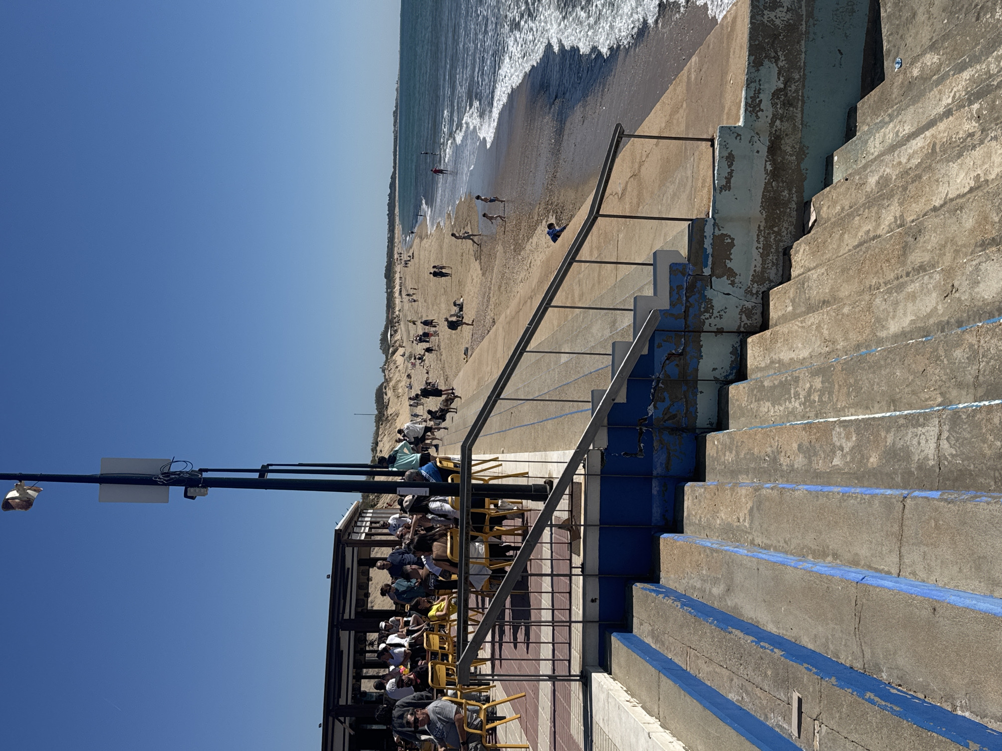 Here is a description of the image:
The image shows a view from a raised concrete structure with blue-trimmed steps, overlooking a beach in Isla Cristina, Spain. On the beach, there are numerous people engaged in various activities such as walking, playing in the water, and sunbathing. A restaurant or cafe adjacent to the steps is filled with people seated at yellow chairs and tables. The sky is clear and blue. Waves are visible in the sea. A street light is located above the restaurant.