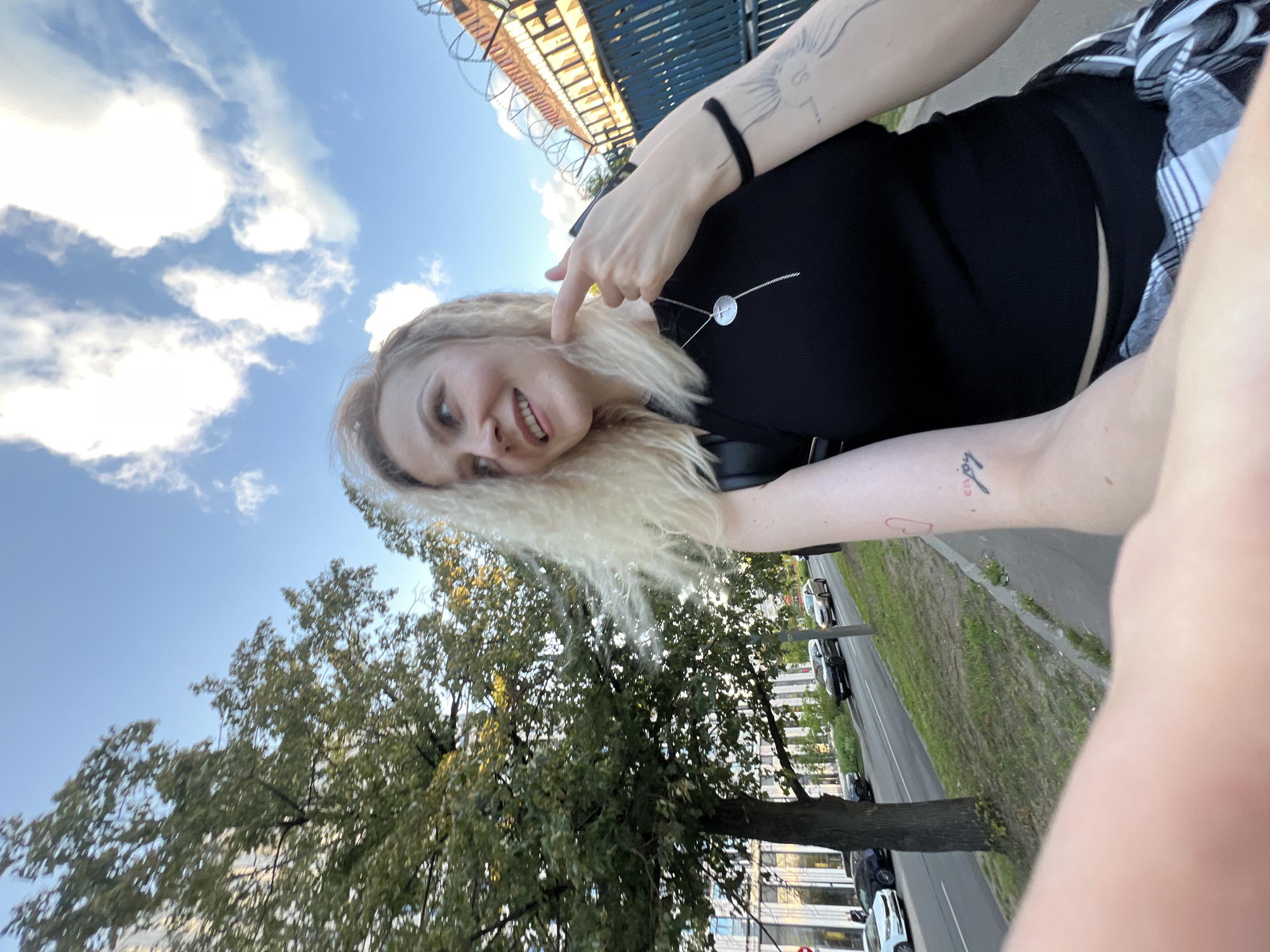 A woman with blonde hair is taking a selfie outdoors. She is wearing a black top and a plaid shirt. She has tattoos on her arms. She is smiling and sticking her tongue out slightly, with her finger pointing towards her cheek.
The background shows a blue sky with white clouds, trees with green leaves, and a road with cars. There are also some buildings visible. The overall scene suggests it is daytime and the weather is pleasant. The presence of a road and buildings indicates an urban or suburban setting.
The angle of the photo and the woman's pose suggest a casual and playful moment.