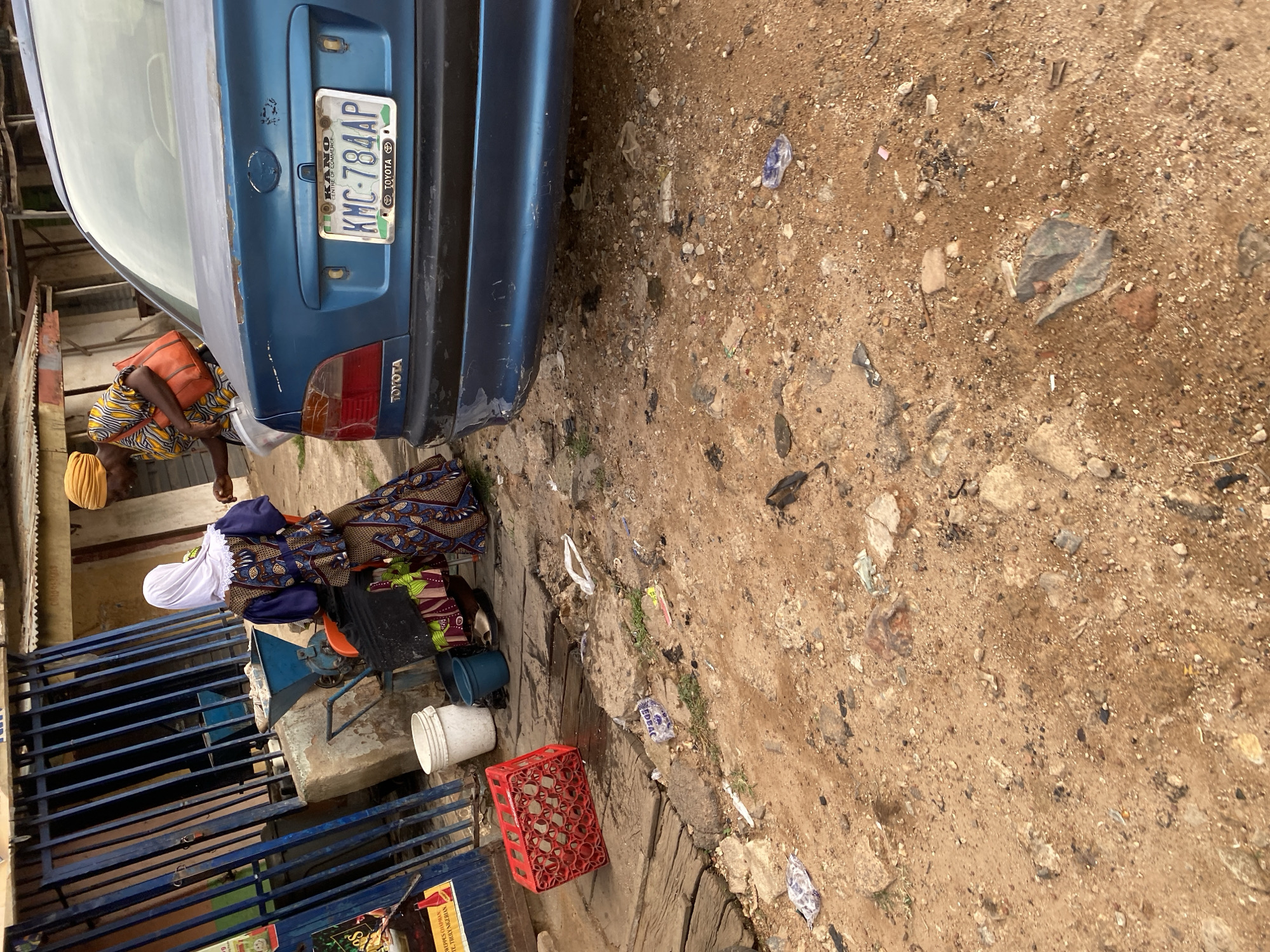 A mid-range shot captures an outdoor scene in what appears to be a market or roadside area in Jos, Nigeria. The foreground is dominated by rough, uneven ground covered in sand, dirt, small stones, and scattered pieces of trash, including plastic wrappers and debris.
To the left, a wooden platform is visible, upon which sits a red plastic crate containing several cylindrical items, possibly bottles or cups. A white plastic bucket and a blue plastic bucket are also present on the platform. Further left, a blue metal gate with vertical bars is seen, with part of a colorful advertisement visible on a stand in front of it. Behind the gate, a concrete structure supports what looks like a grinding machine with a blue funnel.
In the middle ground, two individuals are interacting. One person, wearing a blue top with an ornate patterned design and a white head covering, is partially visible from the back, seated or stooped. They are wearing a patterned skirt and holding a black object. Beside them, another person, wearing a yellow and patterned orange top and a yellow headwrap, is holding a white basin and appears to be engaged in some activity with the first person.
To the right, the rear of a blue Toyota sedan is prominent. The car has a license plate that reads "KANO KMC-784AP" with "CENTRE OF COMMERCE" and "TOYOTA" also visible. The car's rear bumper shows signs of wear and tear. The background consists of a building with a corrugated metal roof and simple structures, typical of roadside businesses. The overall impression is a bustling, somewhat cluttered outdoor commercial environment.