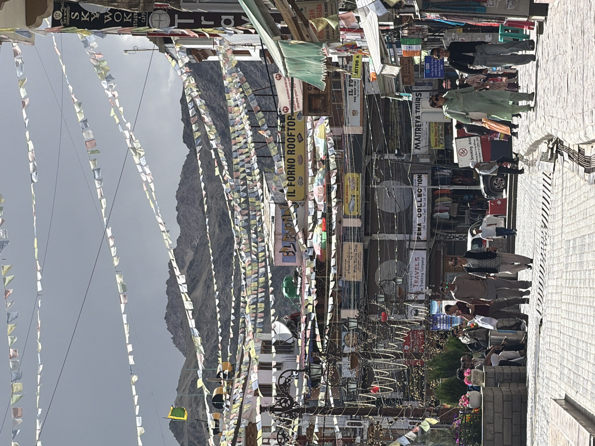 The image depicts a bustling street scene in Leh, India, likely on the main market road. Prayer flags are strung across the street at various levels, creating a colorful canopy against a partly cloudy sky. The street is paved with cobblestones, and on either side, buildings house shops and restaurants, indicated by numerous signs in English and possibly local languages. Some of the visible signs include "SKY WOK RESTAURANT & CAFE," "IL FORNO ROOFTOP," "MAITREYA TOURS," and "TRAVELS THE TREKKING COMPANY."
People are seen walking on the street and in front of shops. In the foreground, a group of individuals are gathered near some potted plants and stone planters. Further down the street, more pedestrians are visible, some shopping or socializing. A white car is parked on the side of the road, and other vehicles are partially visible. The architecture appears to be a mix of traditional and more modern styles, with multiple stories and balconies. The overall atmosphere suggests a vibrant commercial area. The mountainous terrain is visible in the background, characteristic of the Leh region.