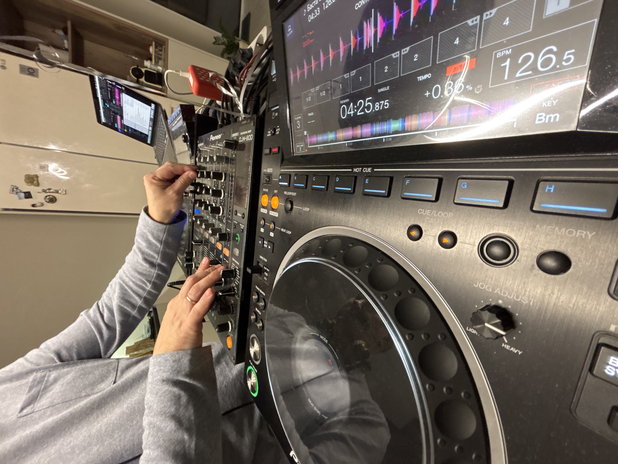 The image shows a person operating a Pioneer DJM-800 DJ mixer and possibly CDJ-3000 media player. The screen on the CDJ-3000 shows a track playing at 126.5 BPM in the key of Bm. The jog wheel adjustment is set to "Ligh" and the track duration is at 04:25.875. A laptop is open behind the mixer. A white LG refrigerator is visible in the background. The location is implied to be in Прикамский, Russia.