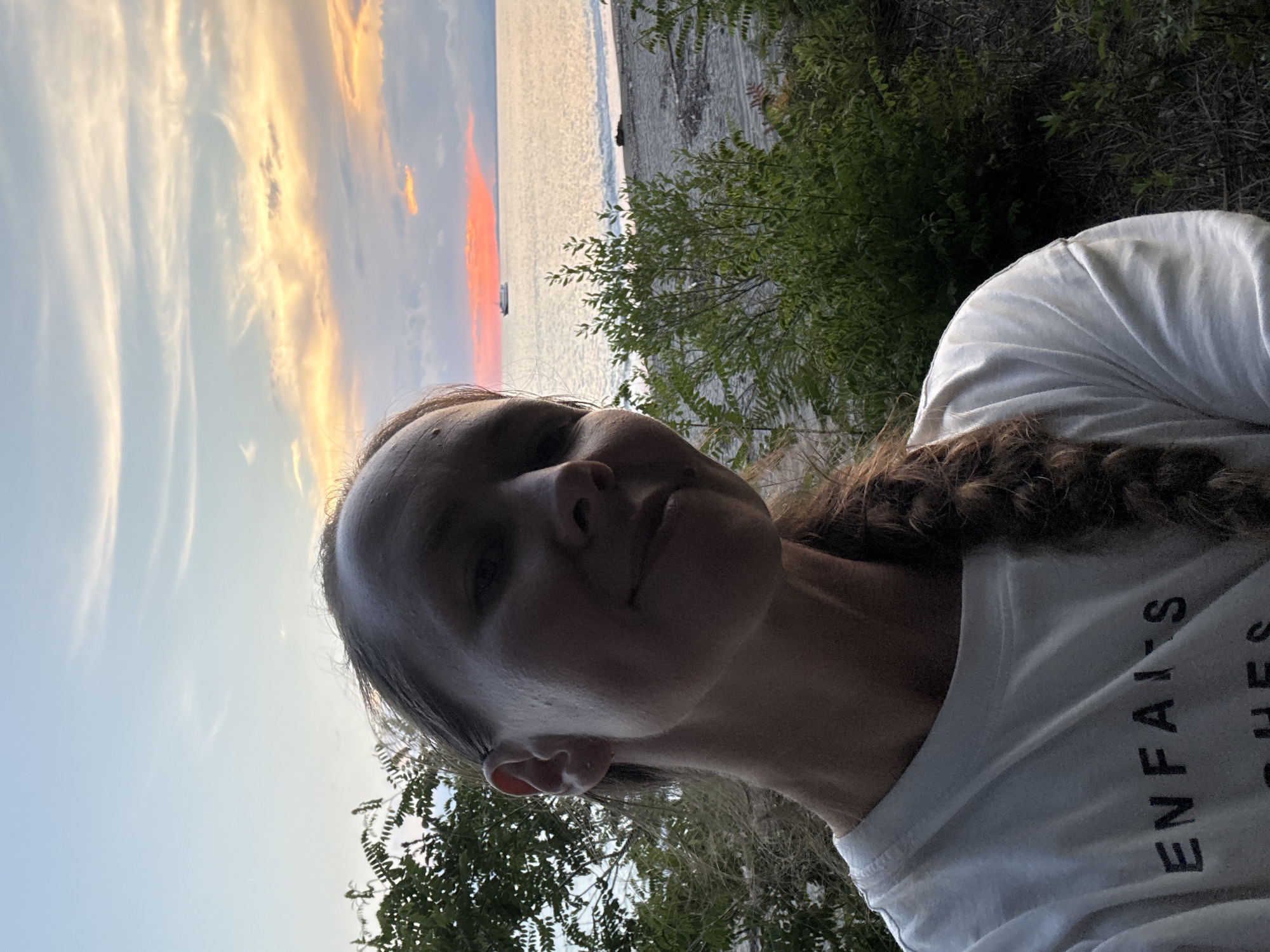 A close-up, over-the-shoulder selfie features a woman with a braid of brown hair, wearing a white t-shirt with black lettering that reads "ENFAITS." She is positioned in the foreground, with lush green foliage to her left and right. In the background, a body of water, likely the sea, is visible, with a beach and small waves. A ship is seen sailing on the water, and the sky is filled with the warm hues of a sunset, featuring pinks, oranges, and blues. The image appears to have been taken outdoors, possibly in the city of Yuada-Gulrypsh, Abkhazia, during the evening.